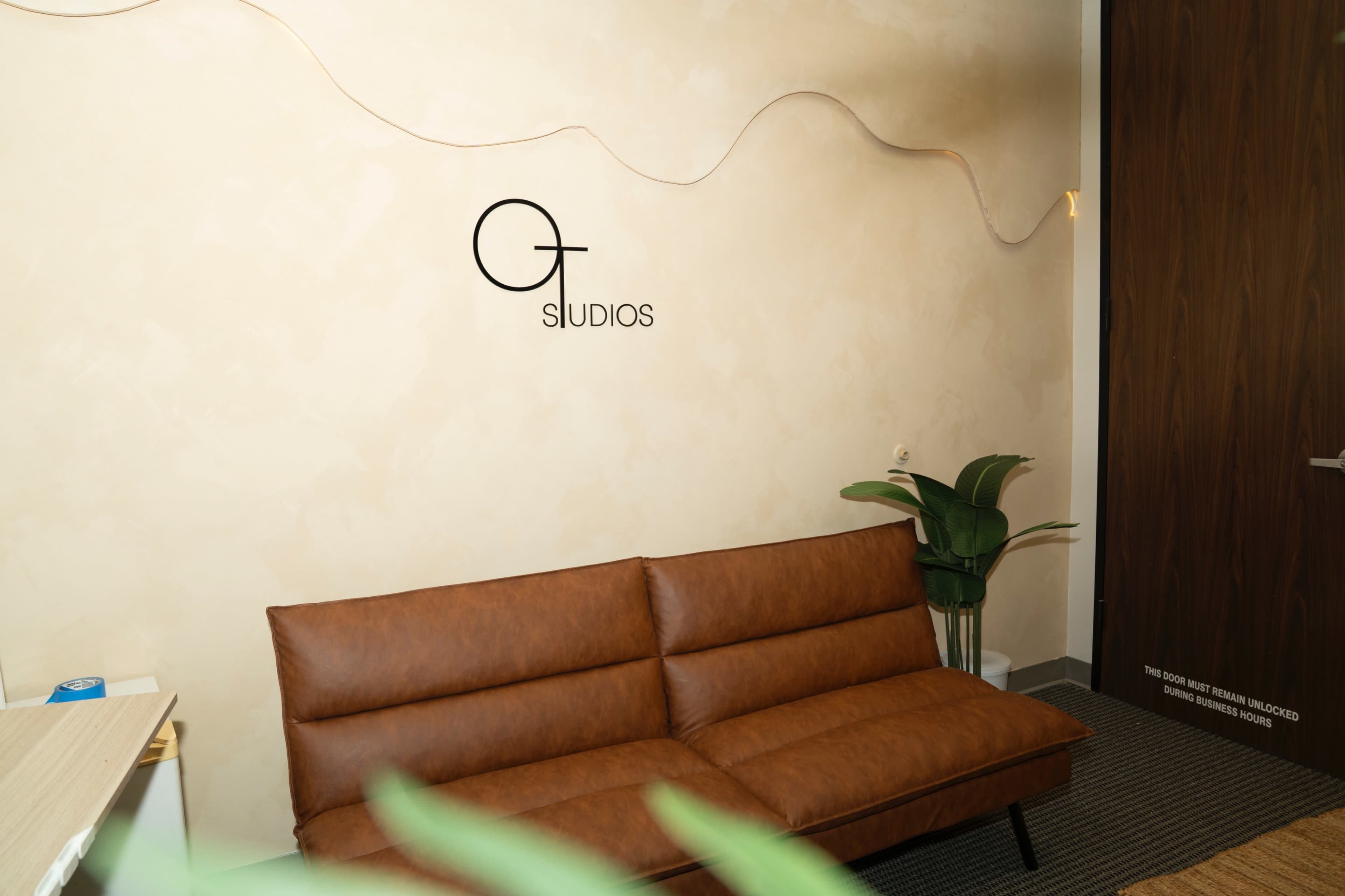 The image shows a modern interior space featuring a brown leather sofa, a potted plant, and a minimalist wall with the word "STUDIOS" displayed on it.