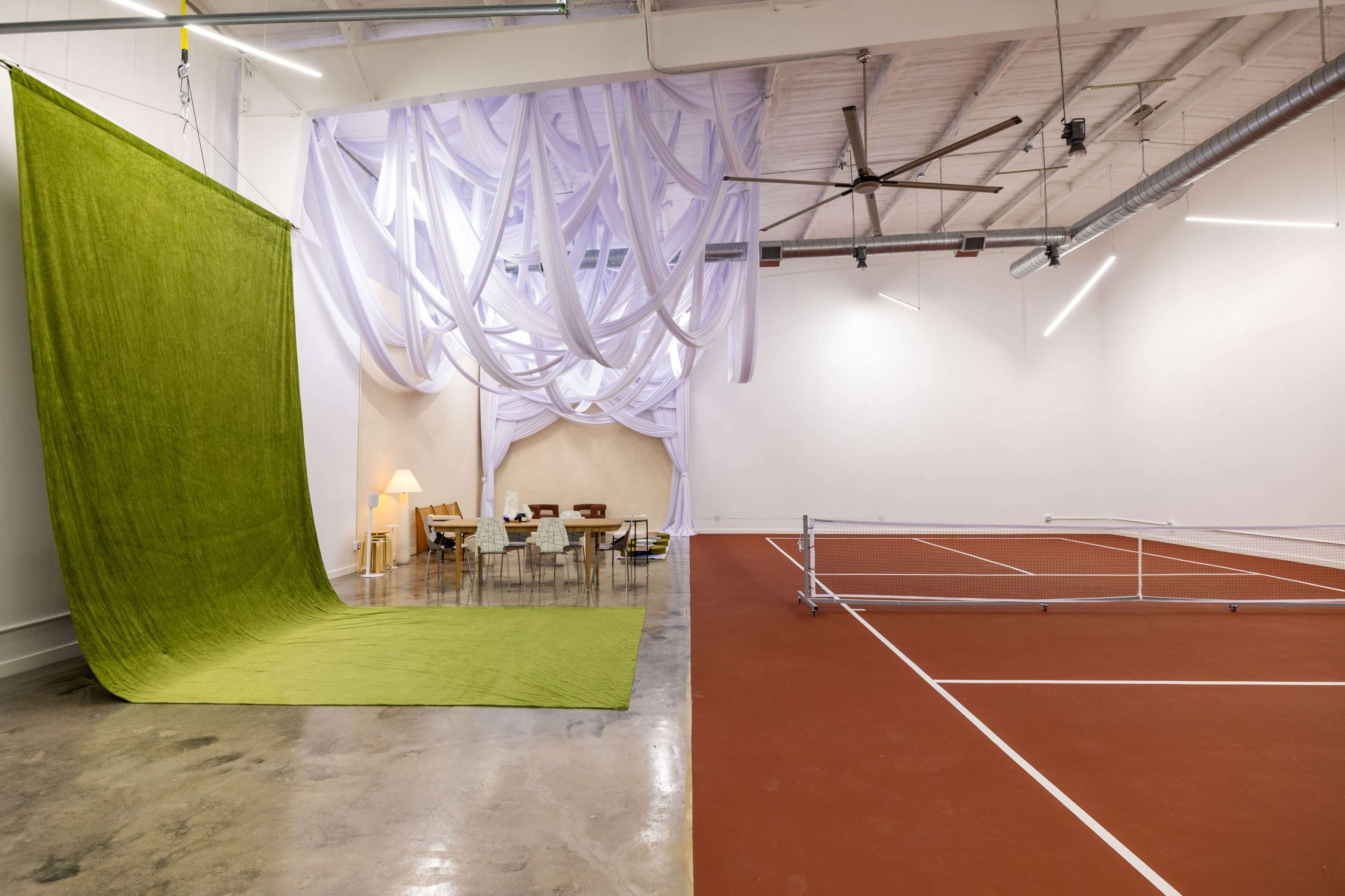 The image shows a spacious room divided by a green fabric backdrop and featuring a tennis court area alongside a seating arrangement with a table and chairs.