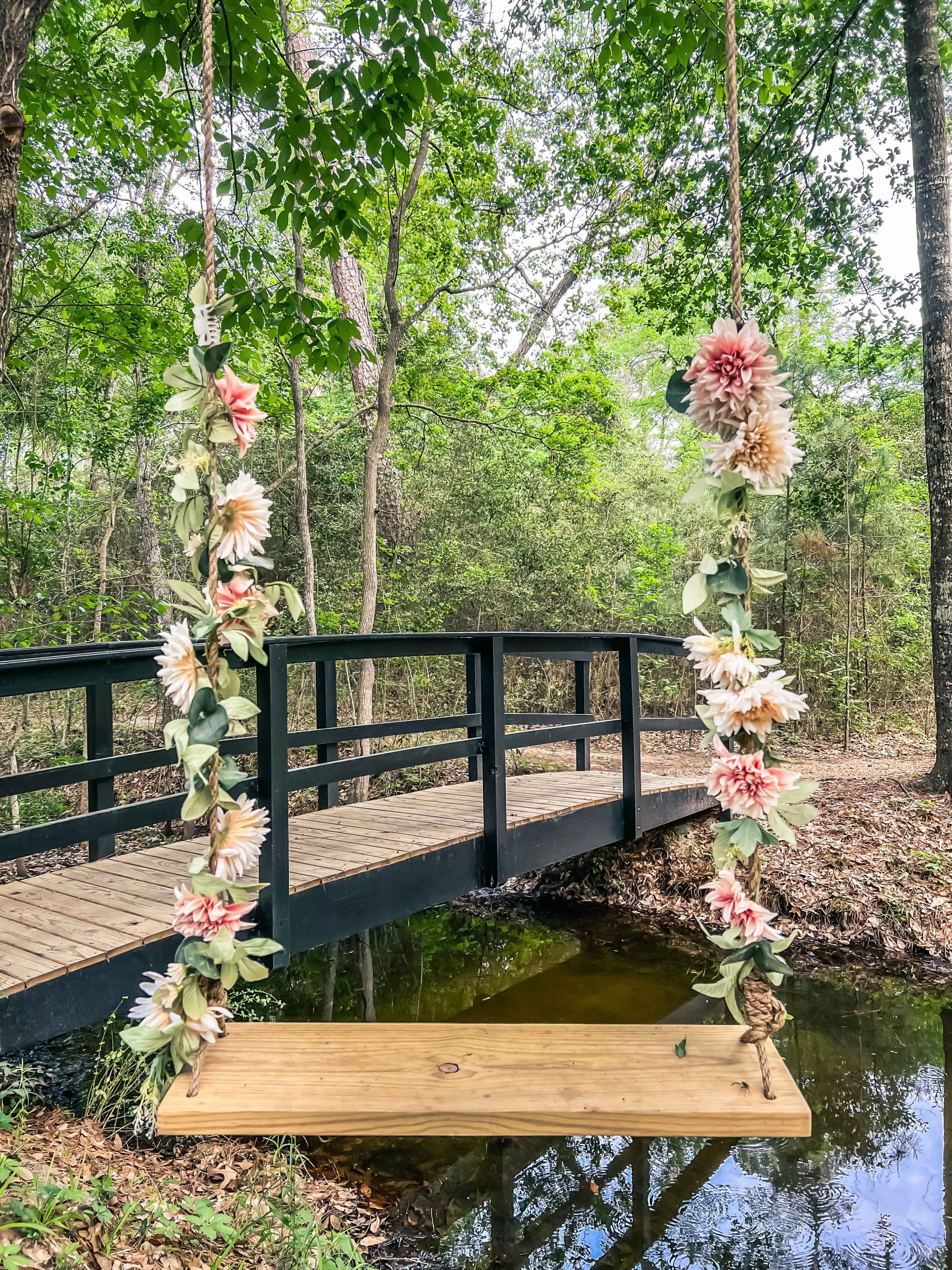 A wooden swing adorned with flowers hangs from a tree near a small bridge crossing over a creek, surrounded by greenery.