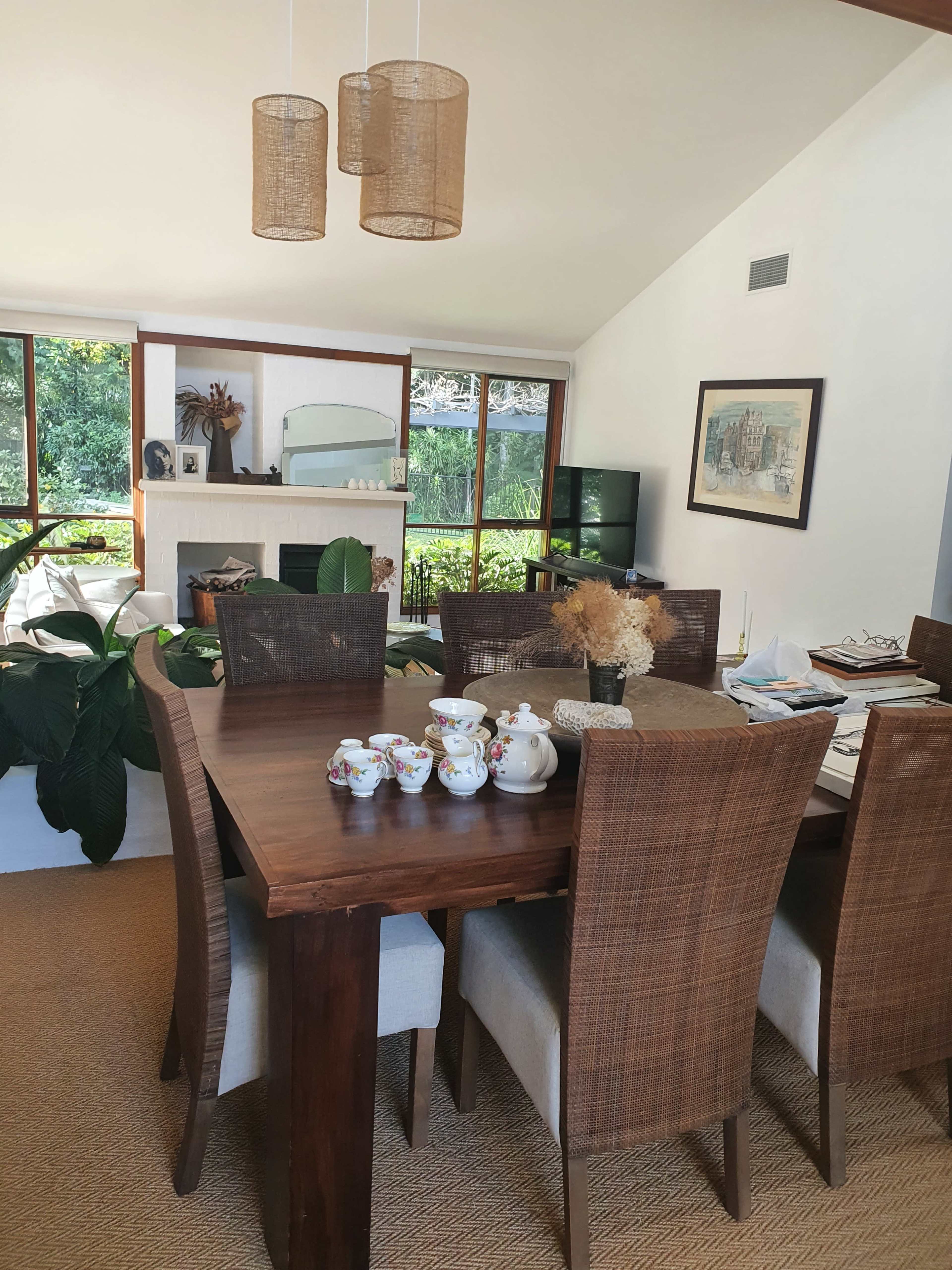 A dining area features a wooden table set with a tea service, surrounded by woven-chairs, with large windows providing a view of lush greenery.