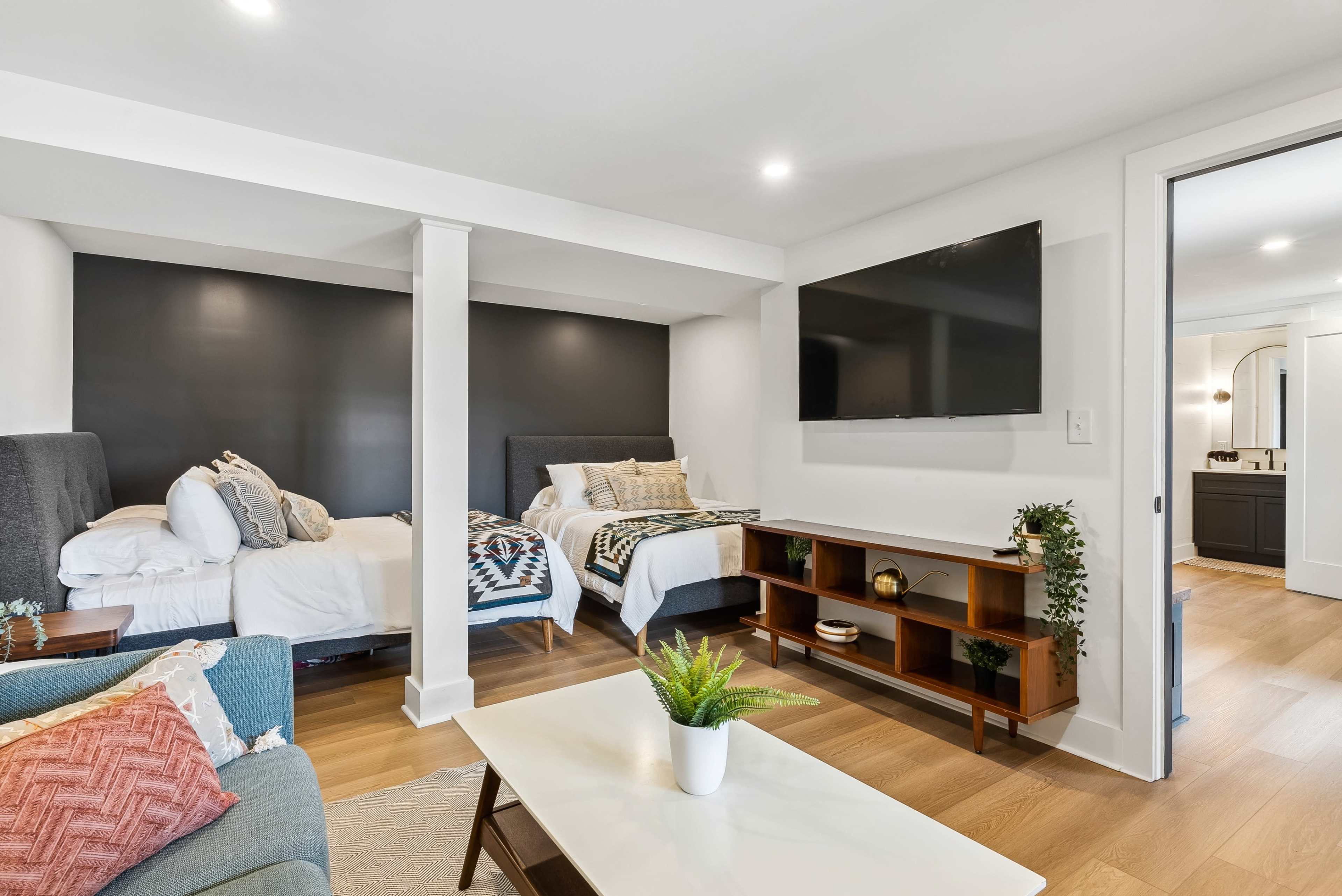 A modern bedroom with two beds, a television mounted on the wall, a coffee table, and a small plant, all set within a well-lit space.