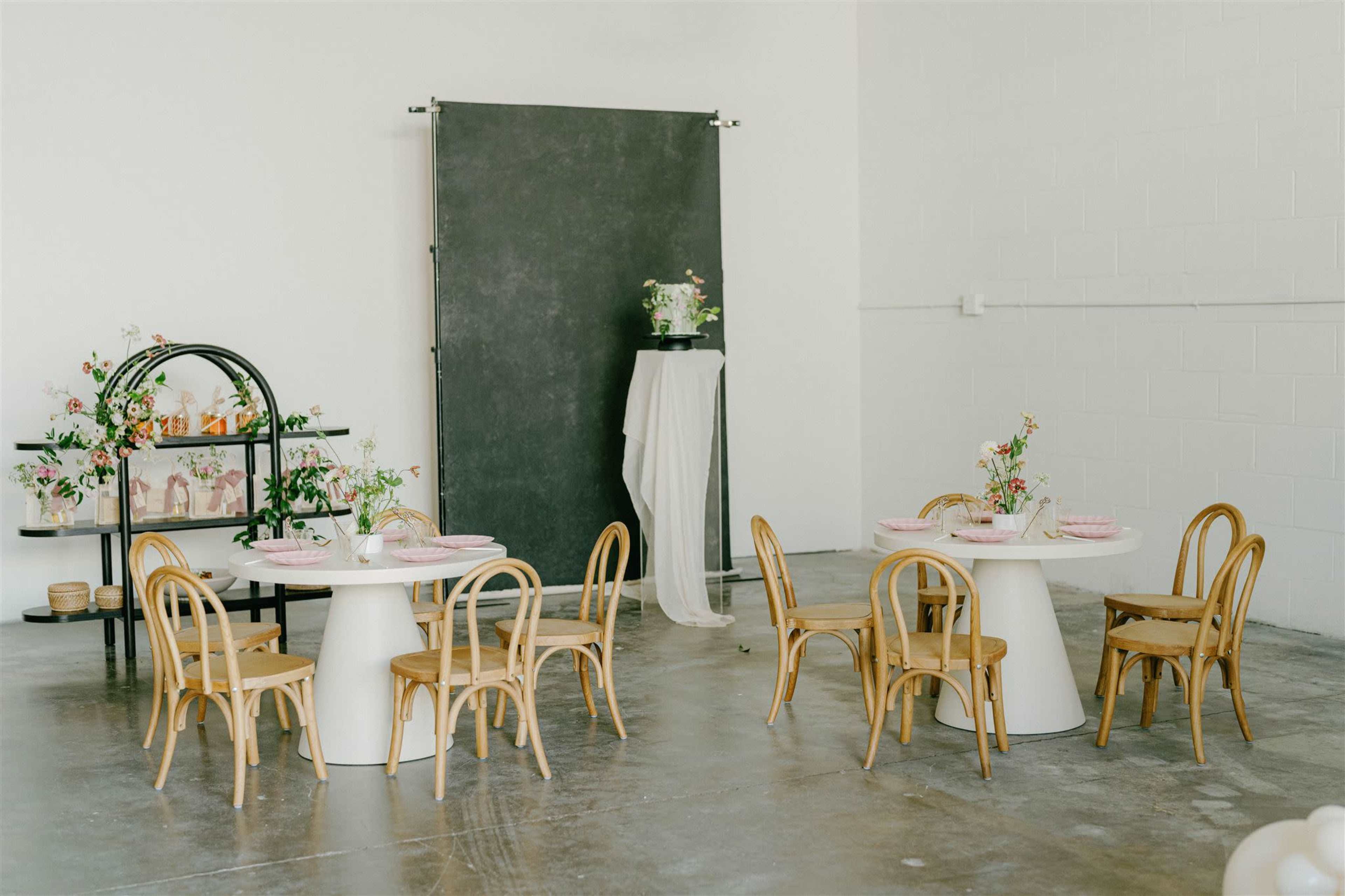 A spacious, well-lit room features two round tables with wooden chairs, a decorative shelf with flowers, and a backdrop made of dark fabric.