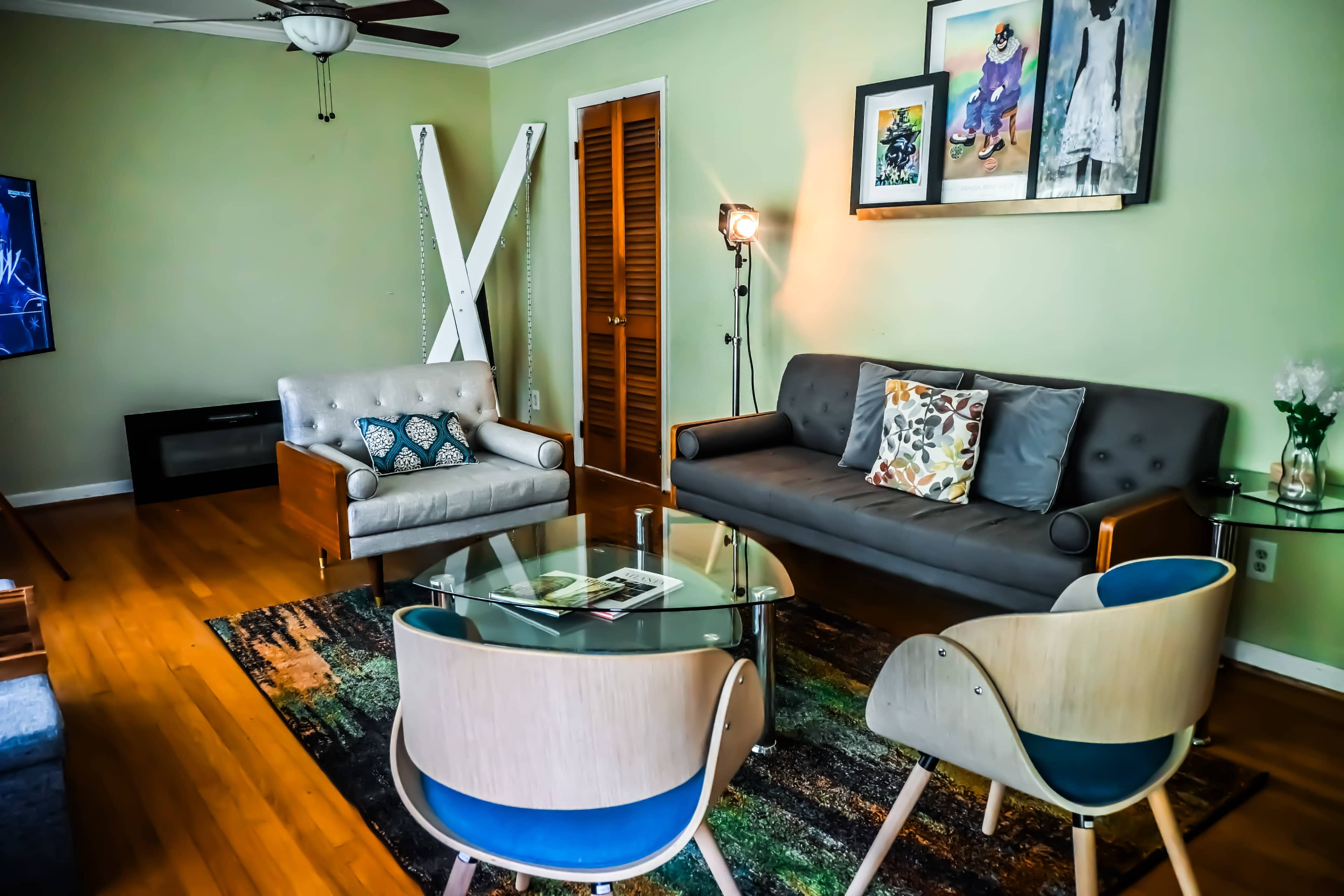 The image shows a modern living room featuring a gray sofa, a light gray armchair, a glass coffee table, and two blue-accented chairs, with a TV in one corner and framed artwork on the walls.