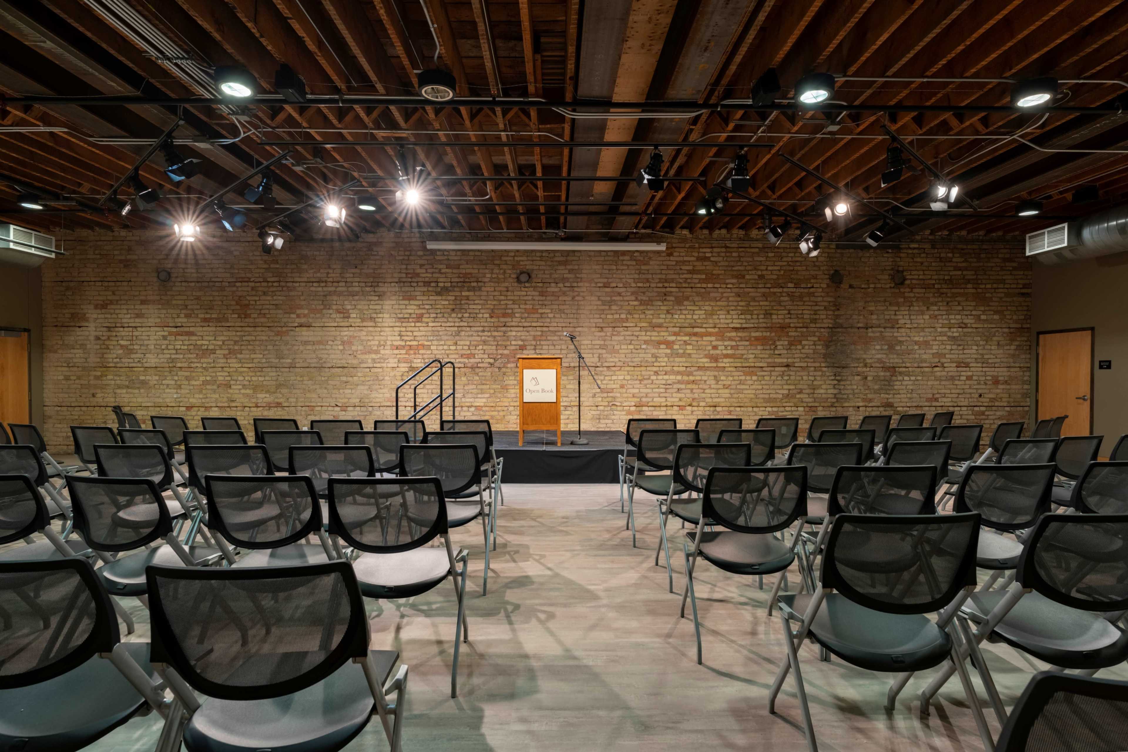 A spacious auditorium features rows of black chairs facing a podium on a stage with a brick wall backdrop and overhead lighting.