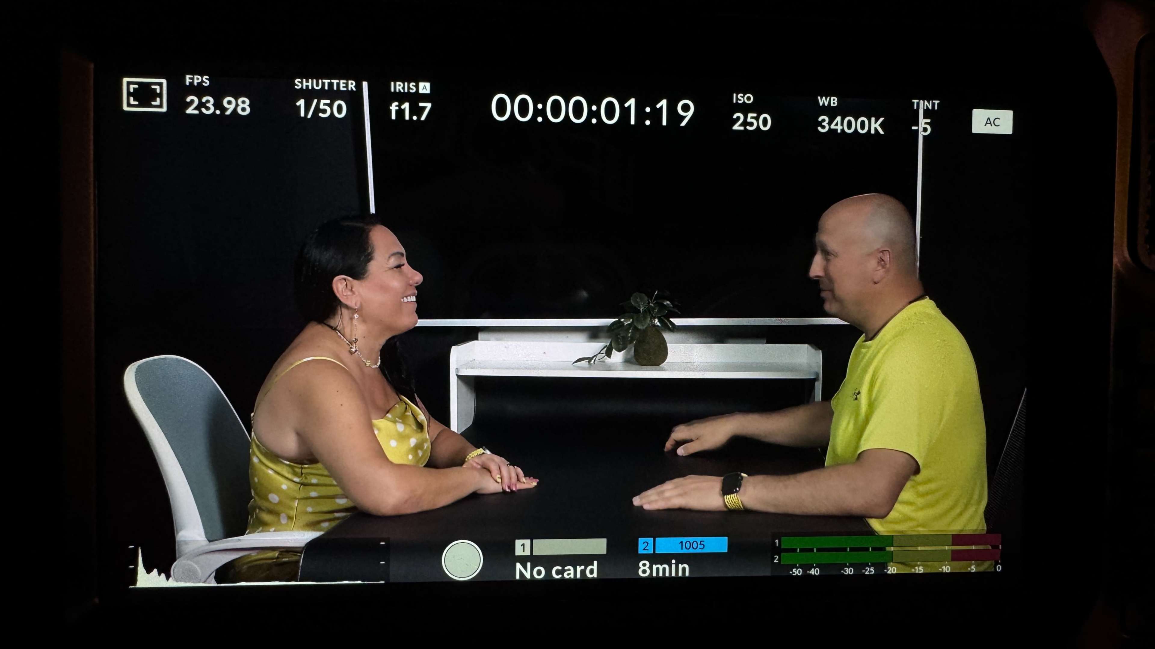 A woman and a man are seated across from each other at a table, engaged in conversation, with a plant and a white shelf in the background.