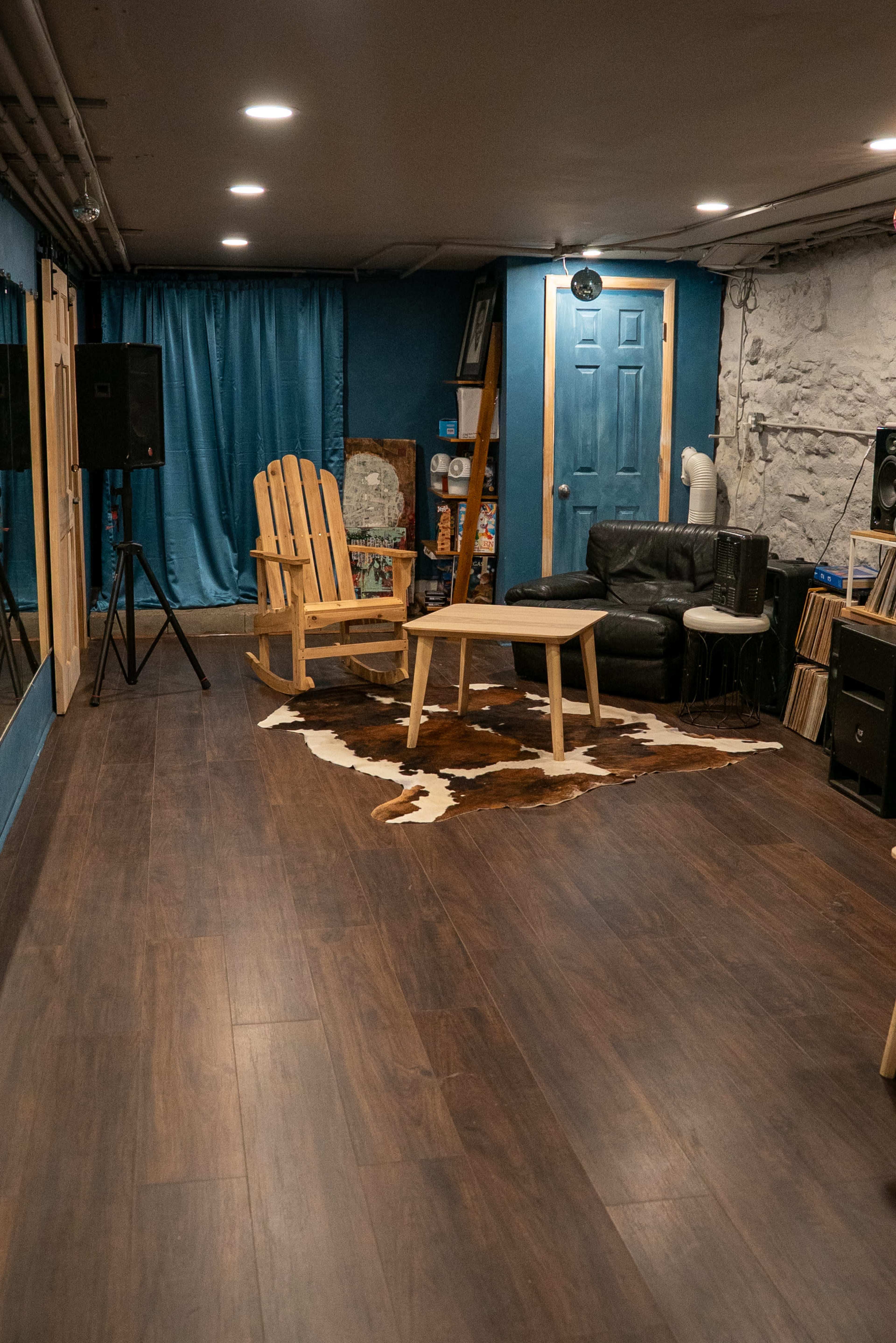 The image shows a spacious basement room with wooden flooring, blue walls, a wooden chair, a small table, and a collection of vinyl records.