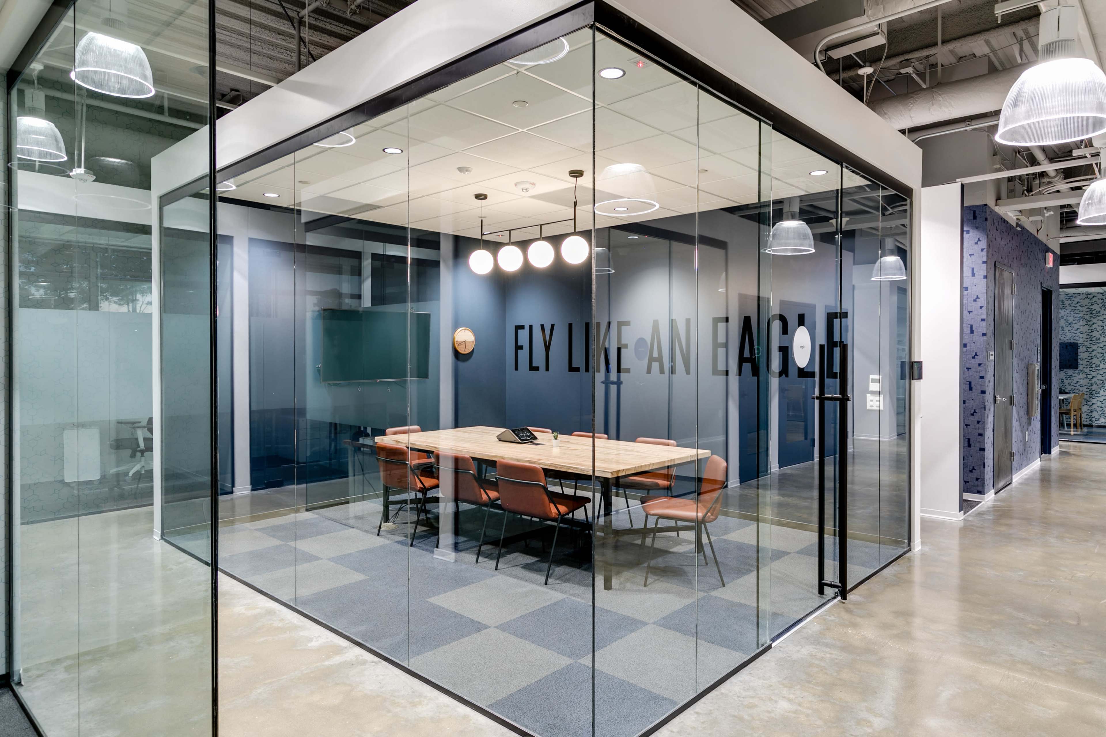 A modern glass-walled conference room contains a large wooden table surrounded by orange chairs, with the phrase "FLY LIKE AN EAGLE" displayed on the wall.