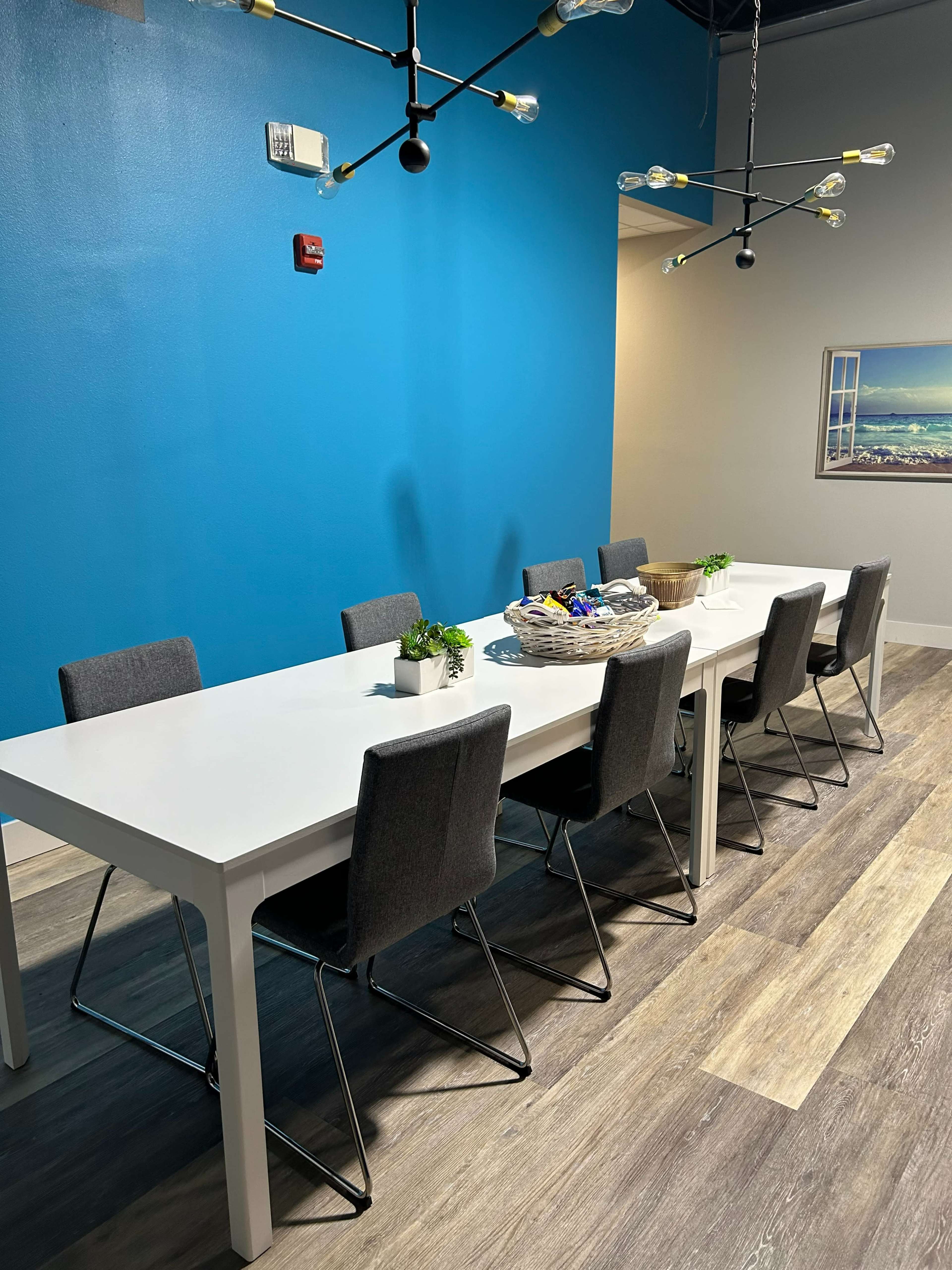 A long, white dining table surrounded by gray chairs is set against a blue wall, with a decorative basket and small plants on the table.