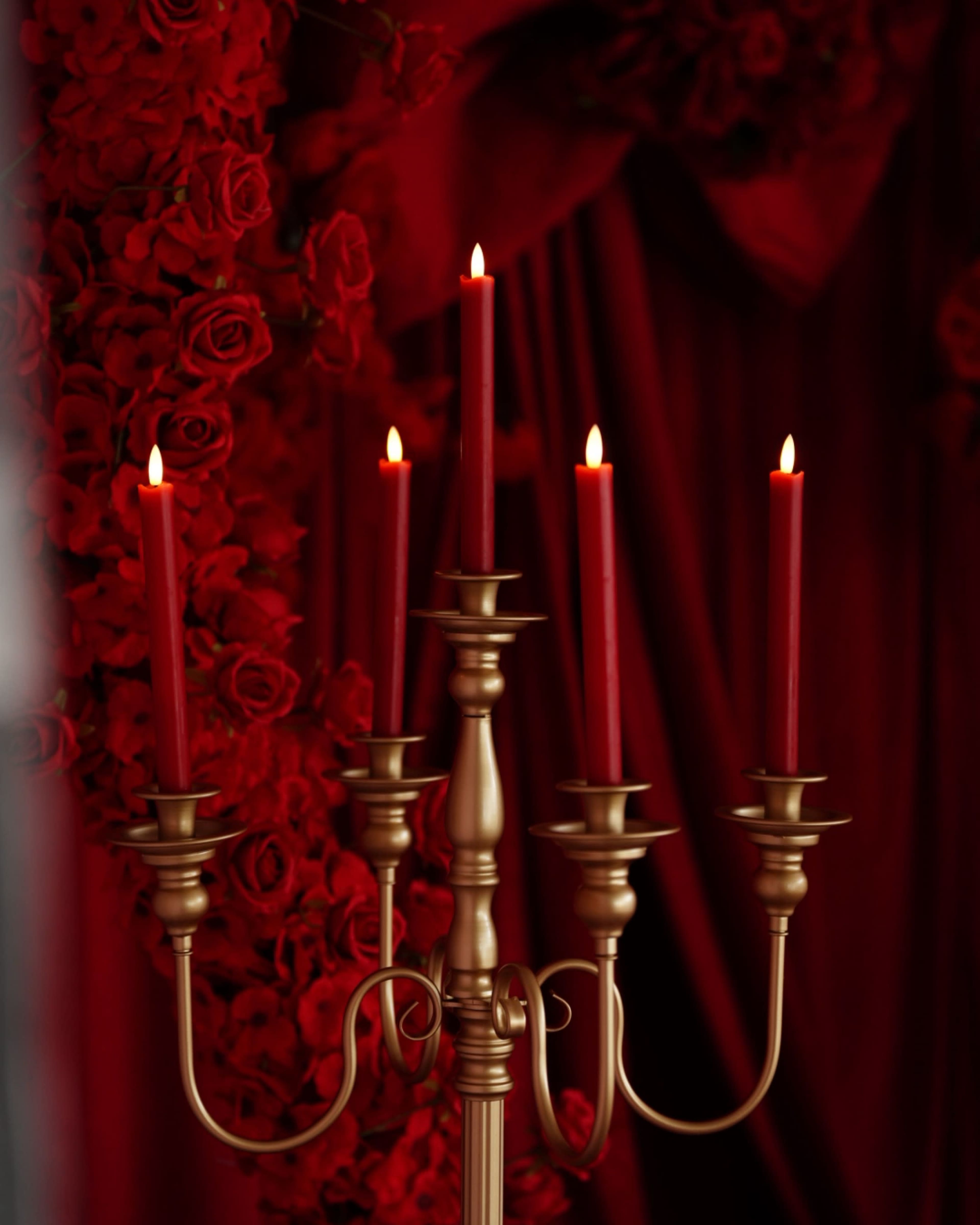 A gold candelabra holds five red candles, set against a backdrop of red roses and fabric.