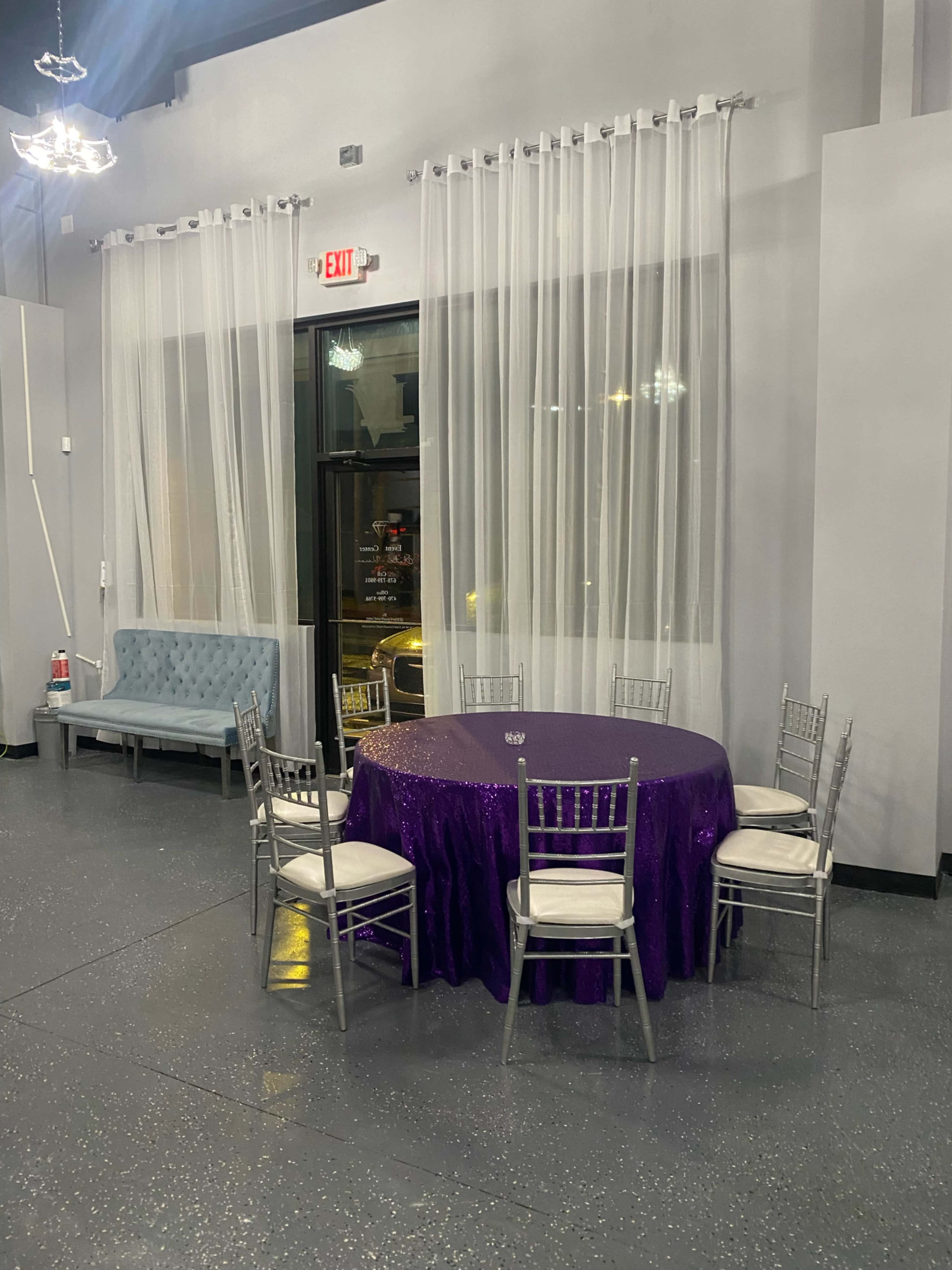 A round table covered with a purple tablecloth and surrounded by silver chairs is placed near a large window with sheer curtains in a well-lit room.