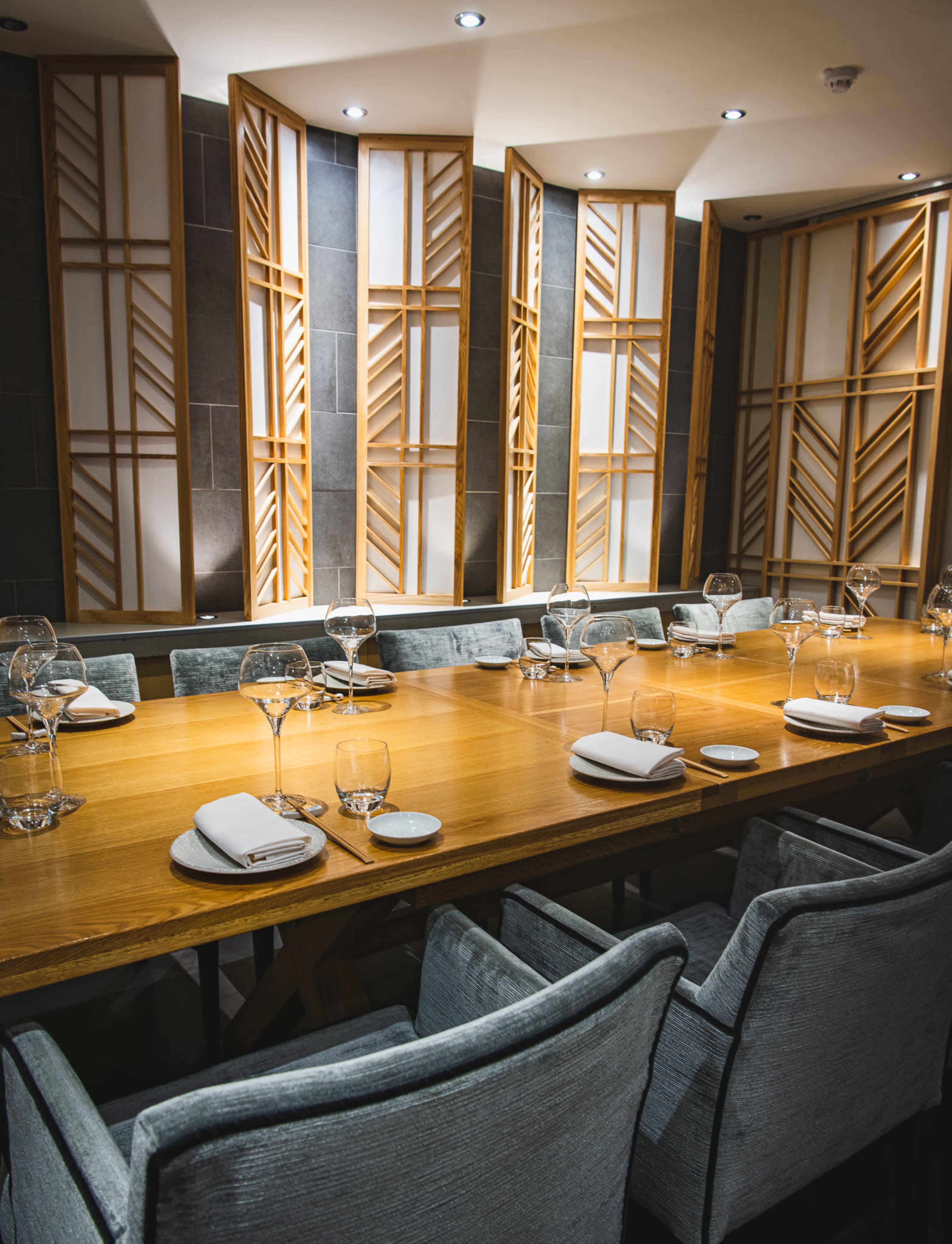 A long wooden dining table is set with glassware and napkins in a modern restaurant featuring wooden wall designs and soft lighting.