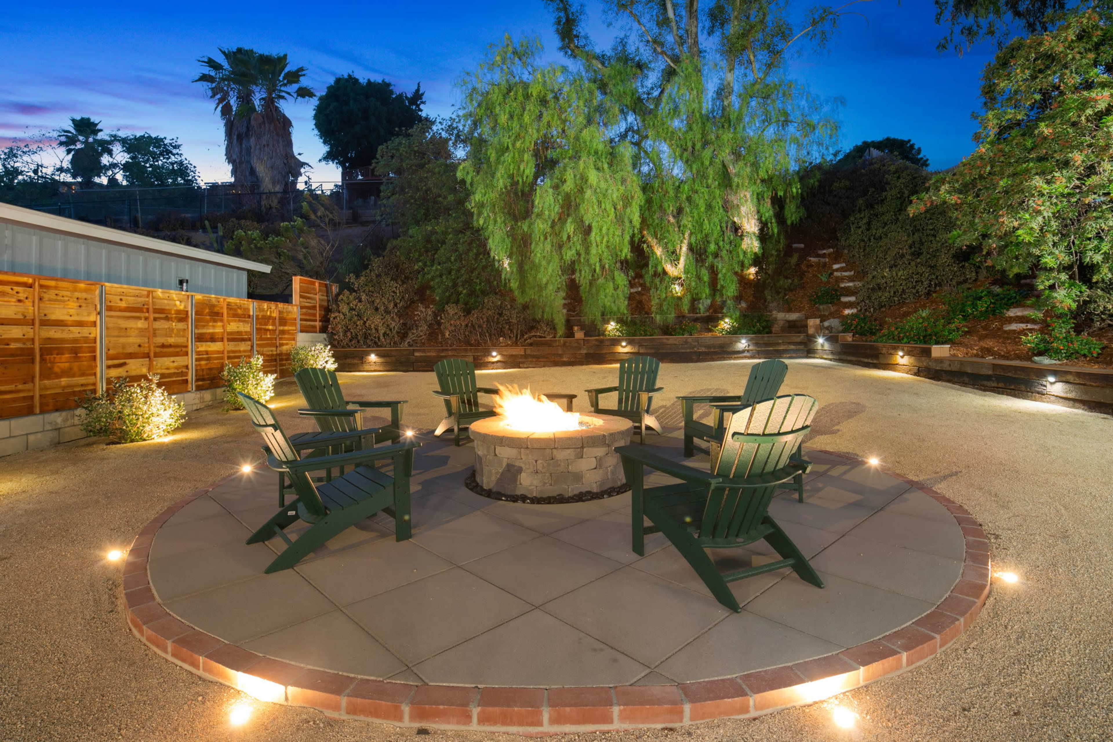 The image shows a circular fire pit surrounded by four green Adirondack chairs, set in a landscaped area with gravel and lighting at dusk.