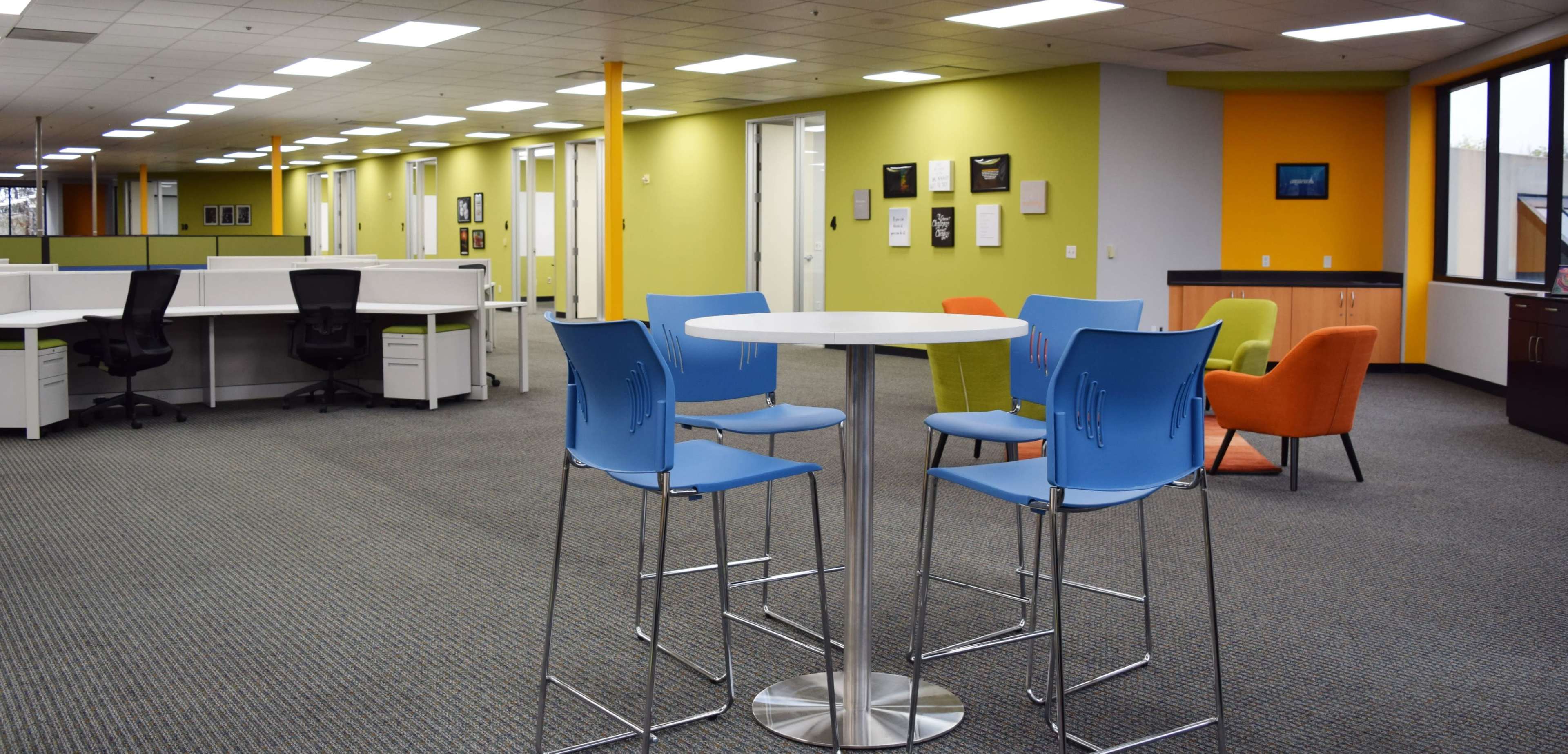 The image shows a modern office space featuring colorful walls, a circular table surrounded by blue chairs, and various workstations in the background.