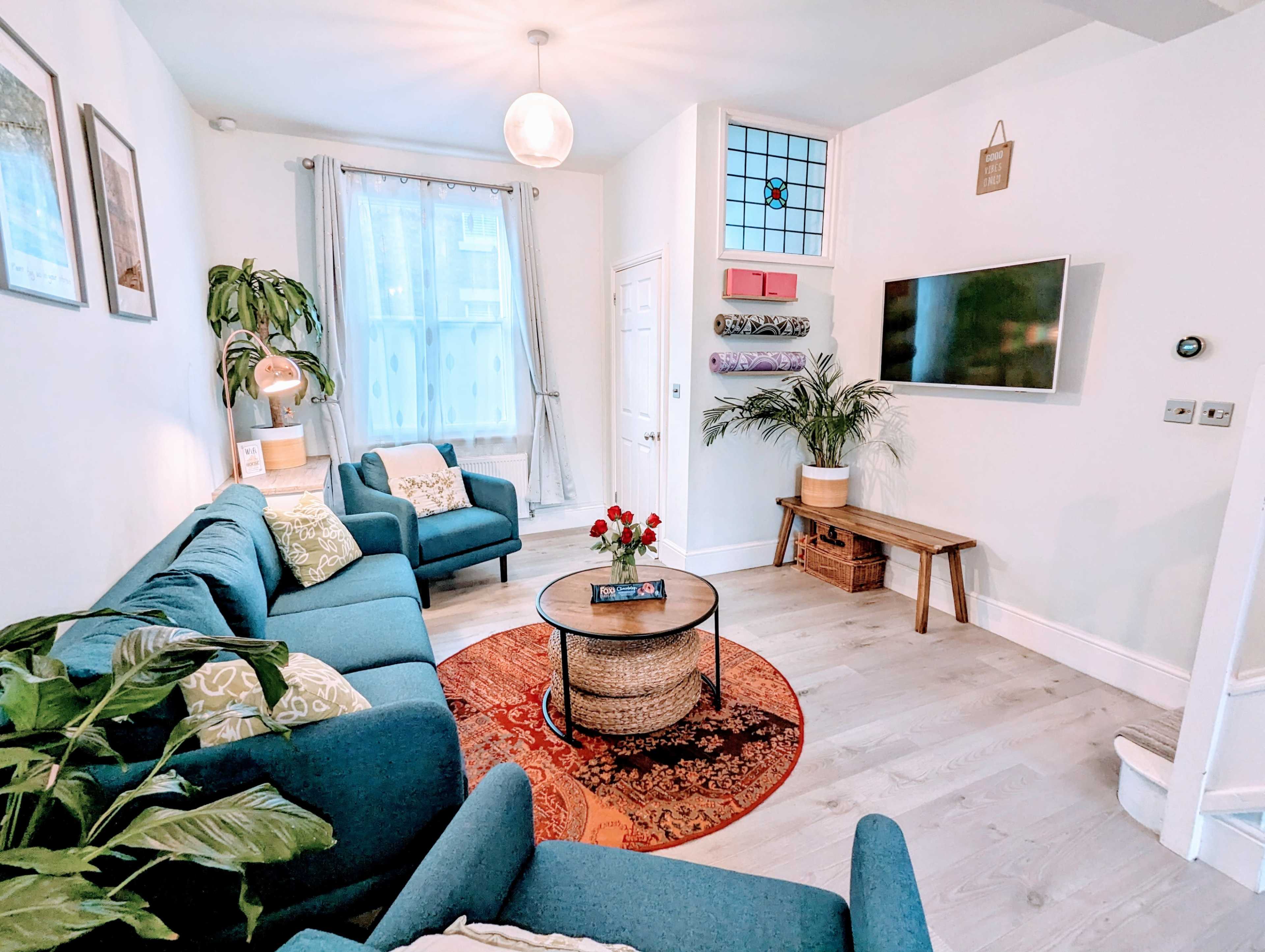 The living room features a blue sectional sofa, a round coffee table with a floral centerpiece, and a wall-mounted TV against a bright, airy background.