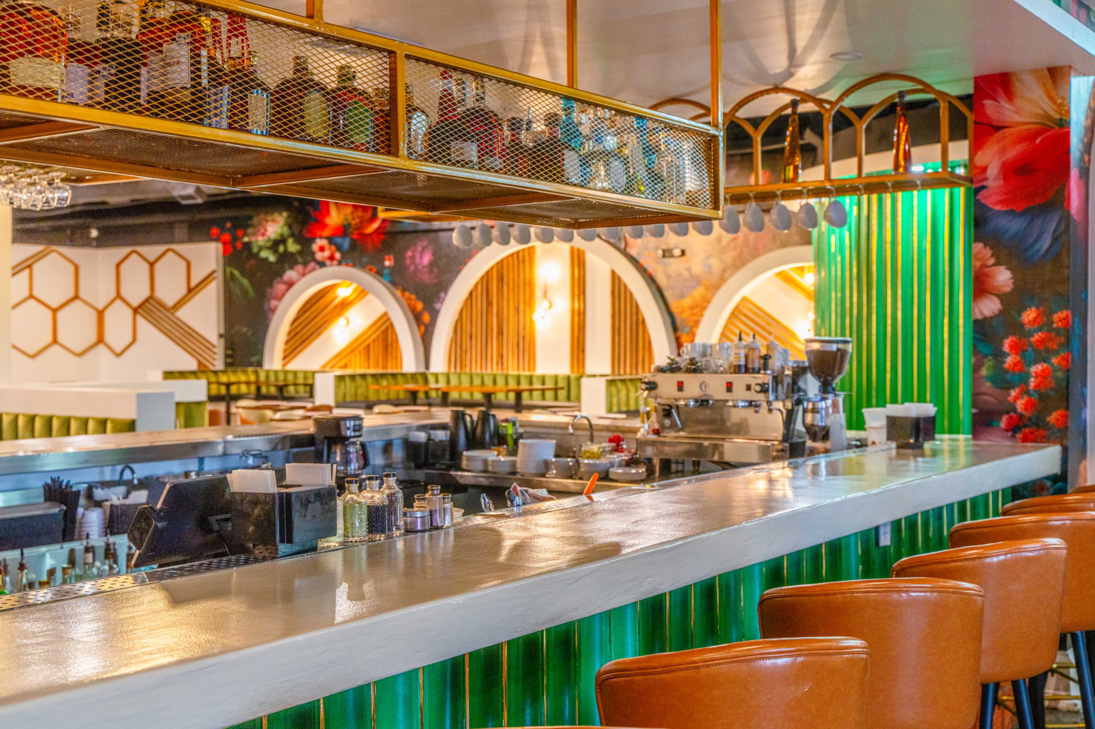 The image shows a modern bar area with a green-striped counter, a coffee machine, and a decorative floral mural in the background.