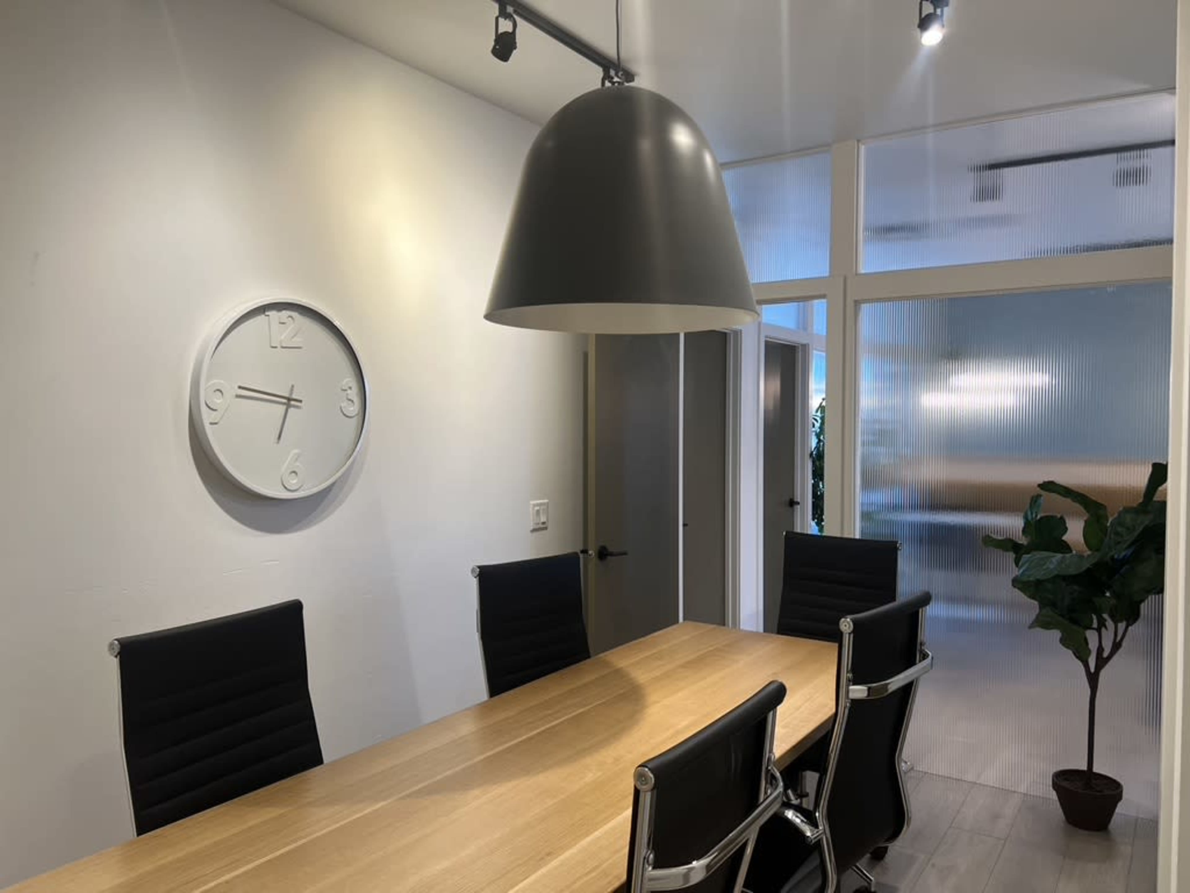 The image shows a modern conference room with a wooden table, black ergonomic chairs, a large pendant light, and a wall clock.