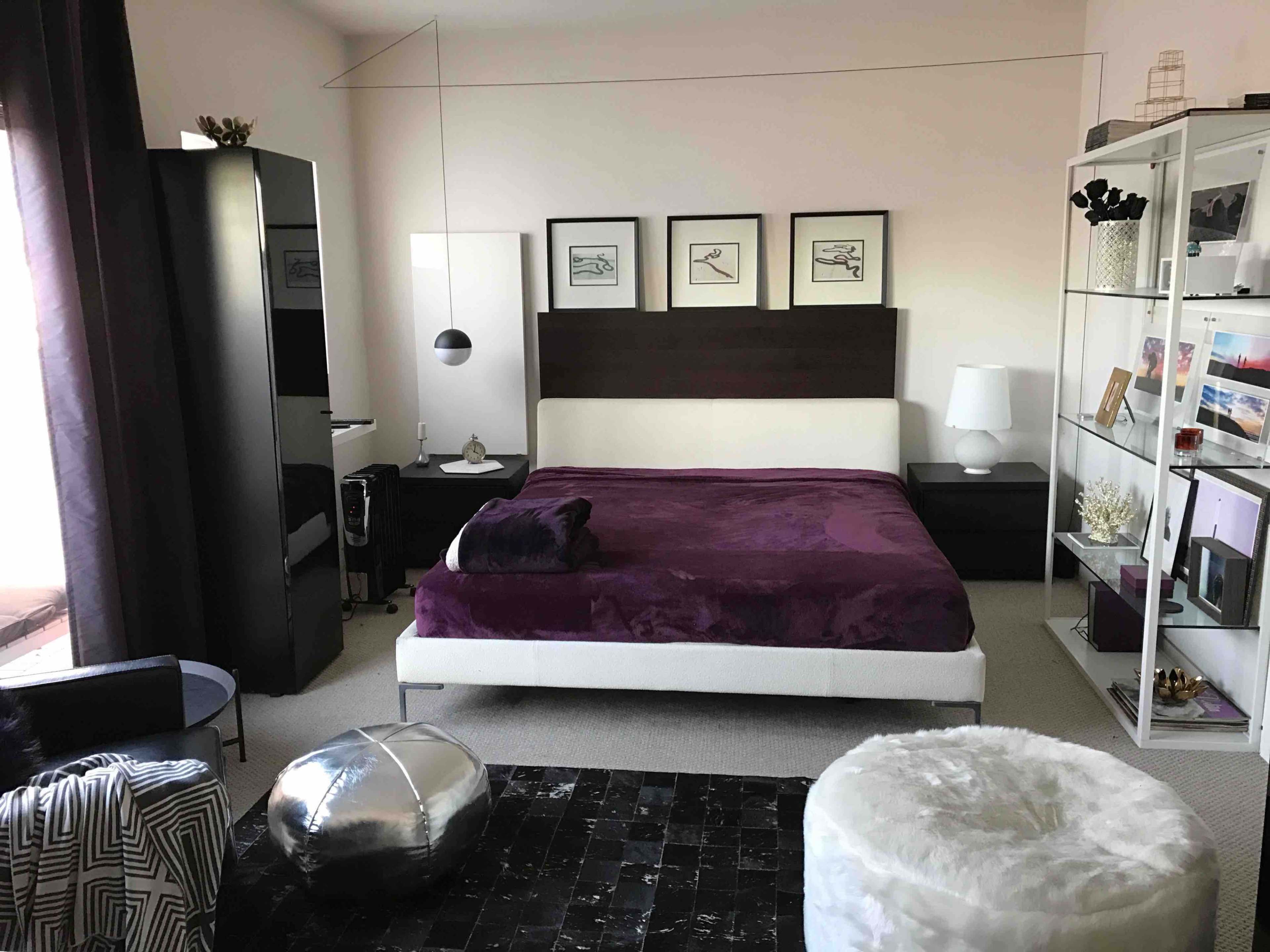 A modern bedroom features a large bed with a purple blanket, framed artwork on the walls, and a combination of dark wood and glass furniture.