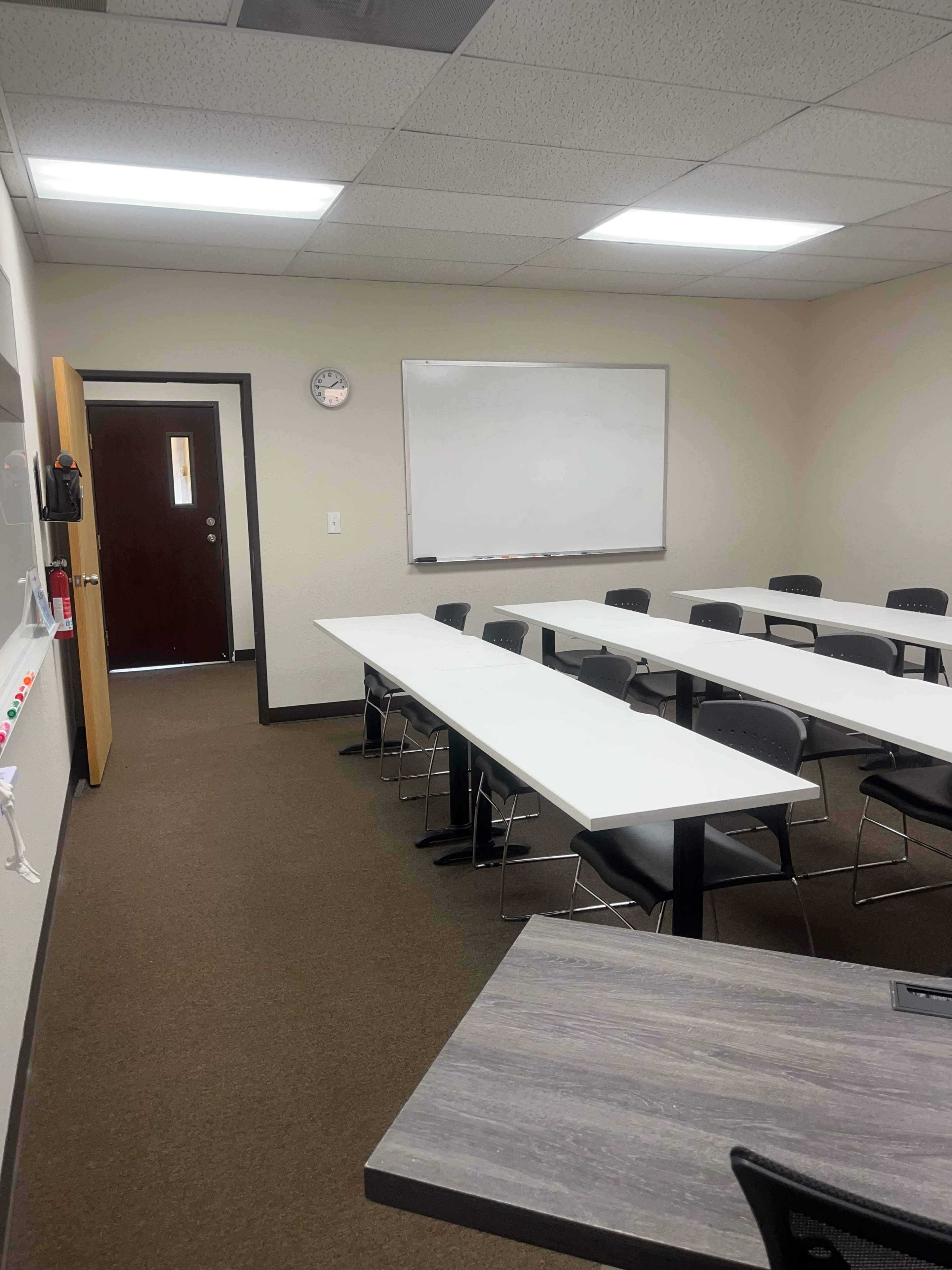 The image shows a classroom with several rows of tables and black chairs, a whiteboard on the wall, and a door leading to a hallway.