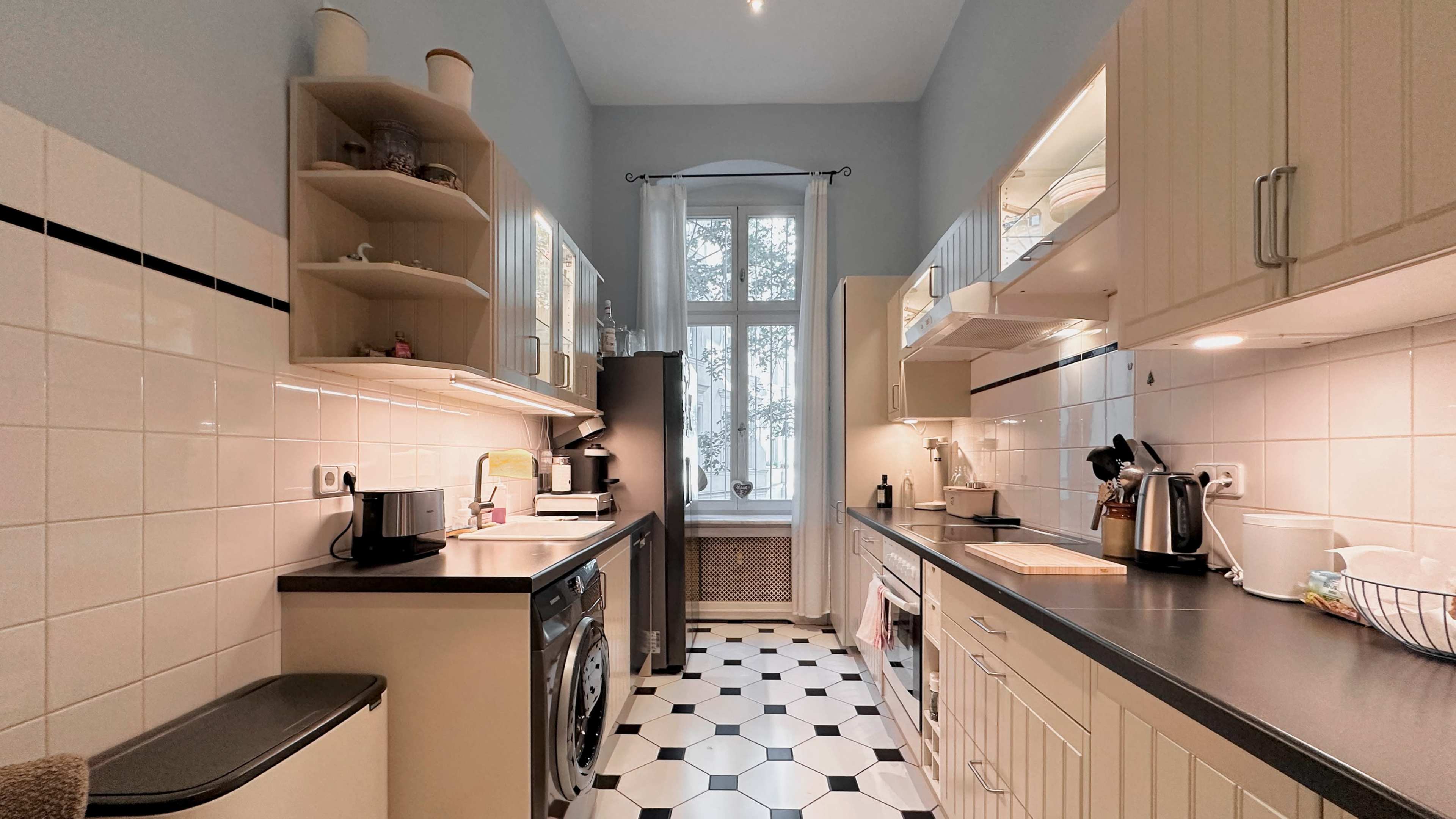 The kitchen features light cabinetry, a washer, and a window that provides natural light, with a patterned black and white tile floor.