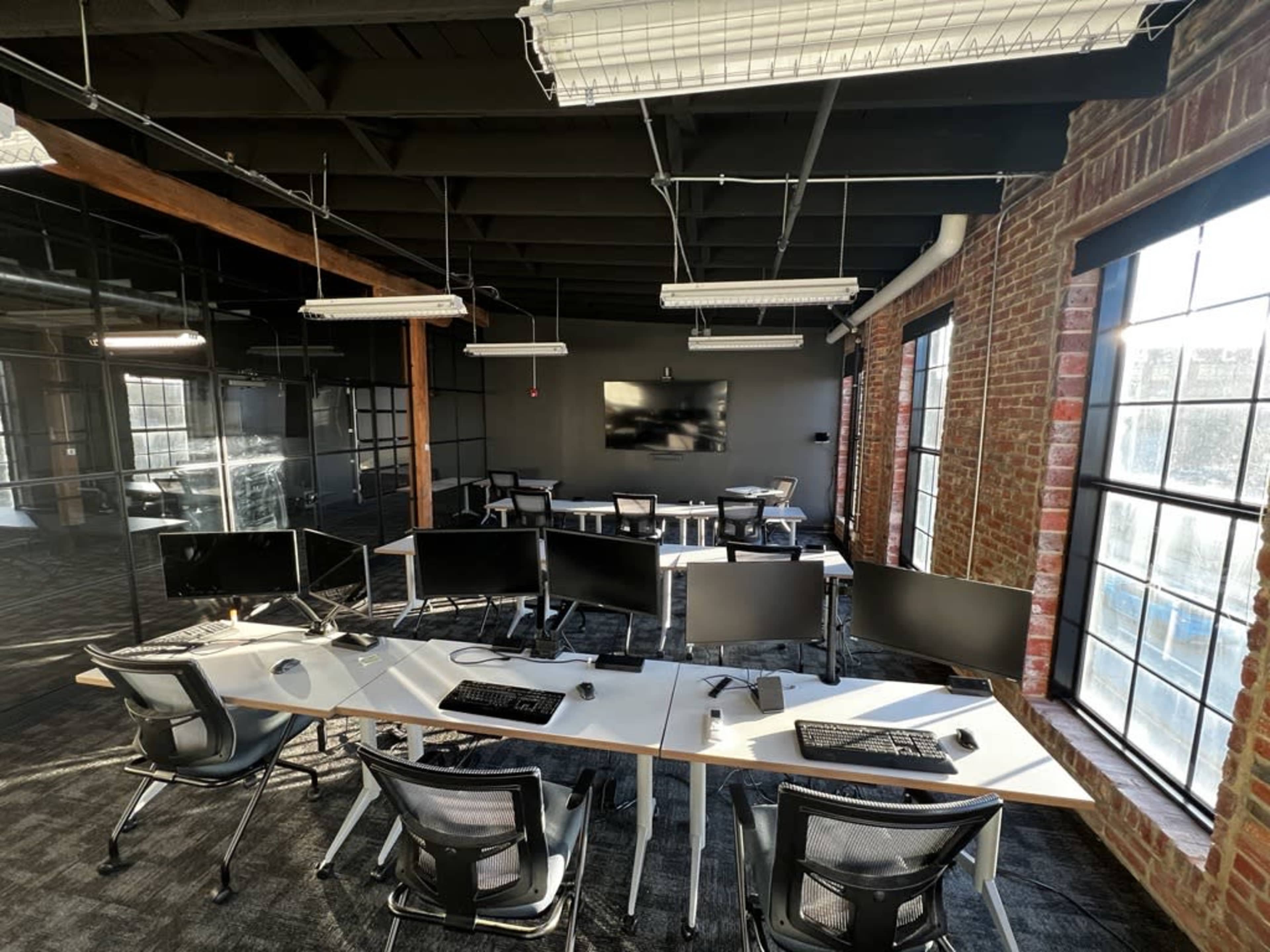 A modern office space featuring multiple computer workstations with monitors, a large screen on the wall, and exposed brick and beam architecture.