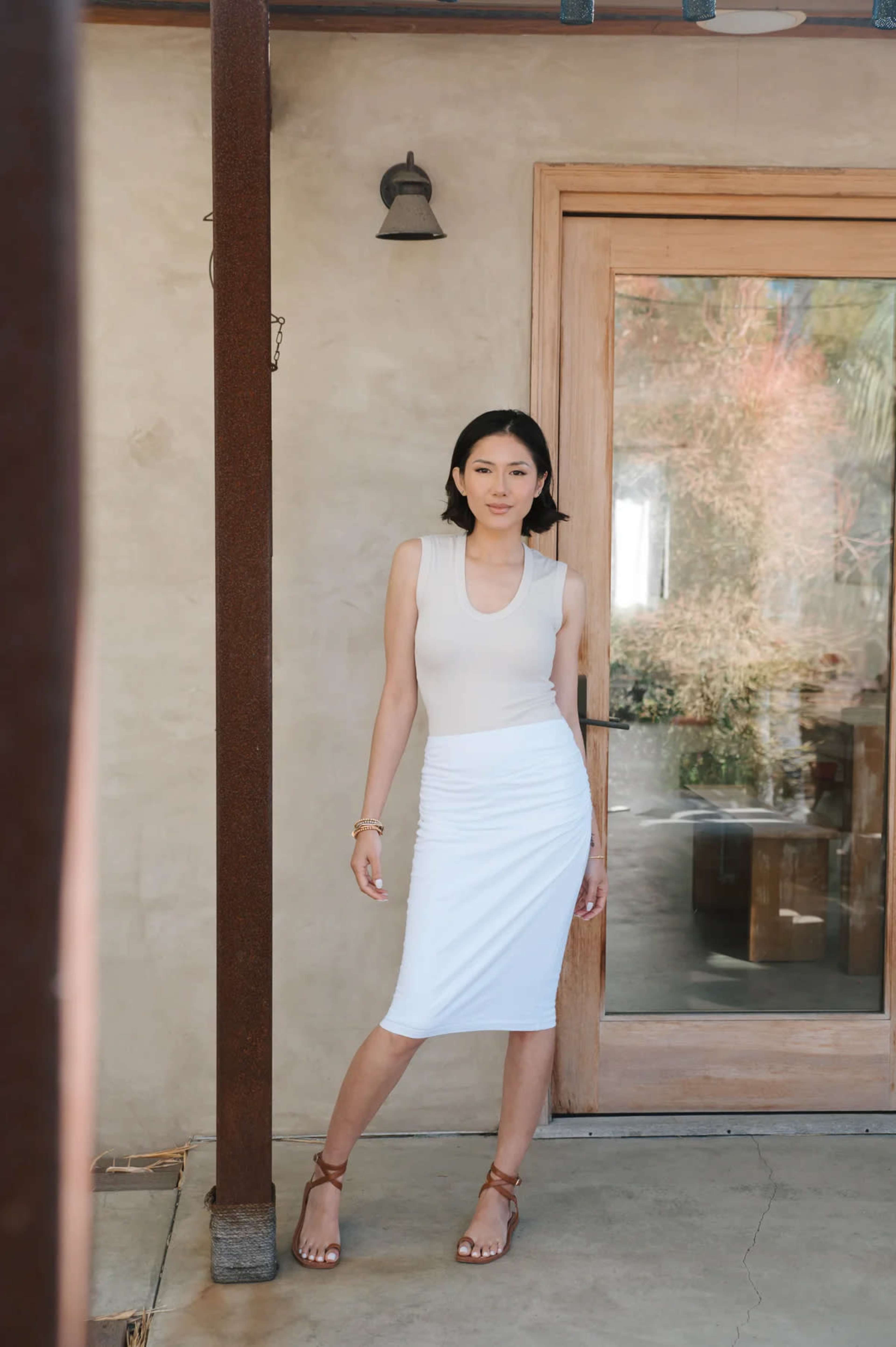 A woman in a fitted sleeveless top and a white midi skirt stands outside a glass door framed by rustic wood.