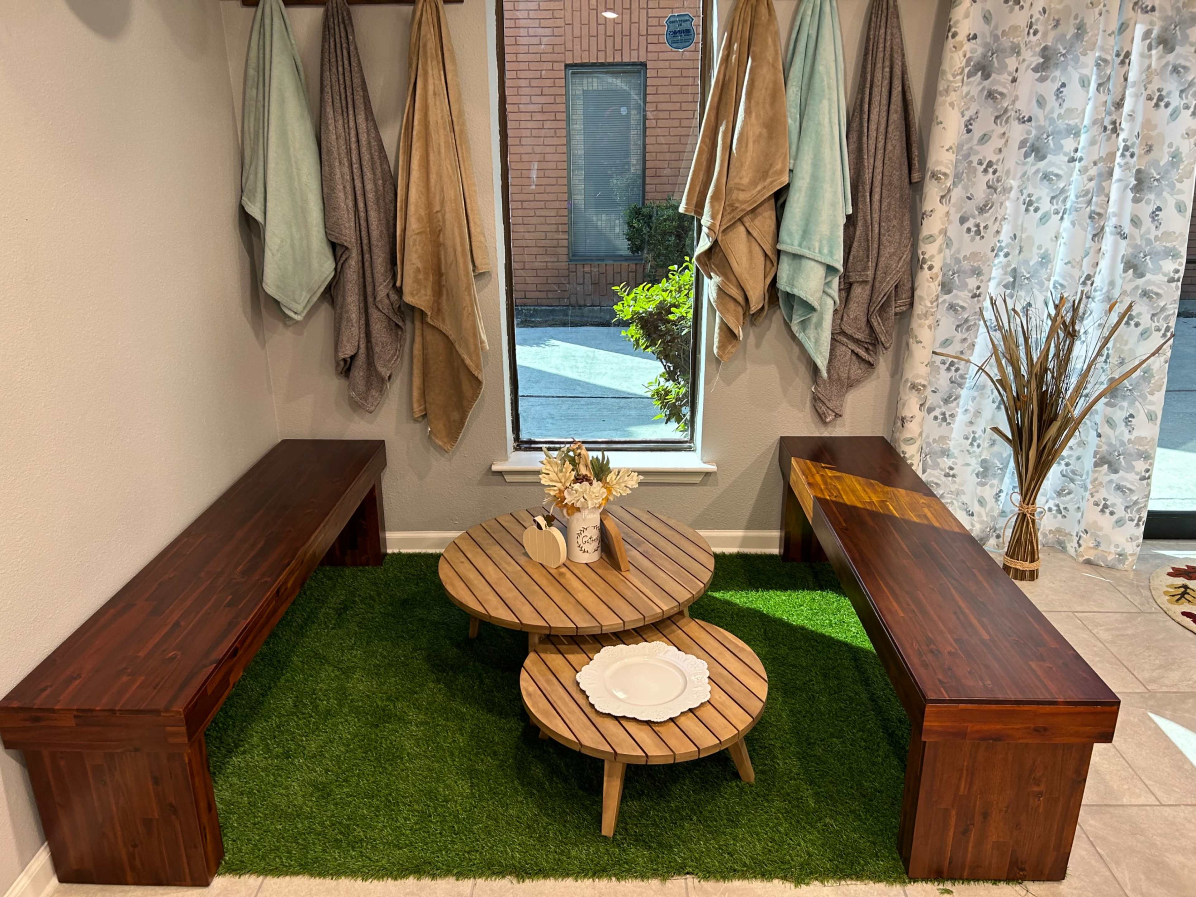 A cozy corner with two wooden benches, a circular wooden coffee table, and decorative plants on green carpet flooring near a large window.