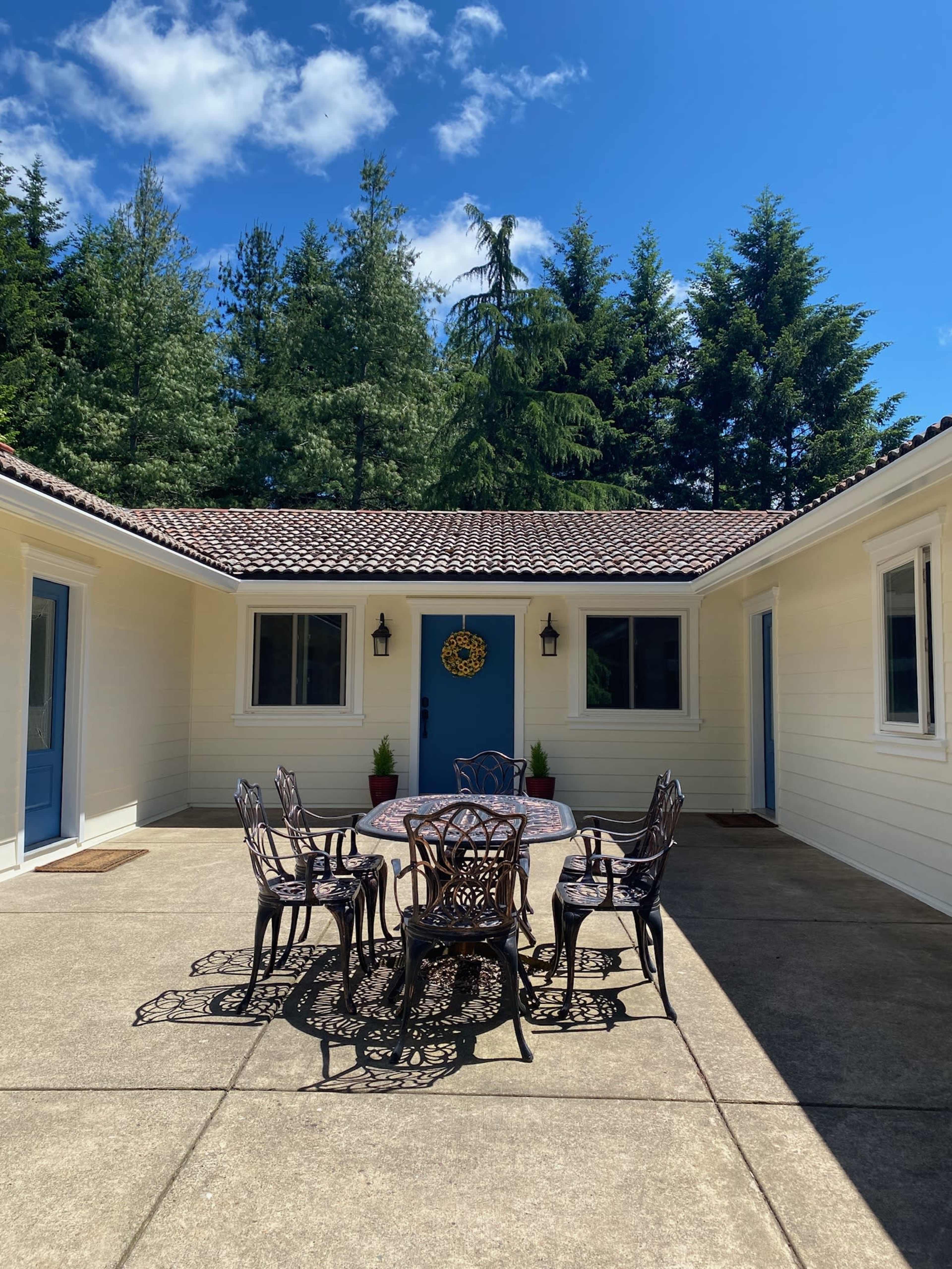 A courtyard features a round table surrounded by six chairs, with a blue door and windows framed by green trees under a clear sky.