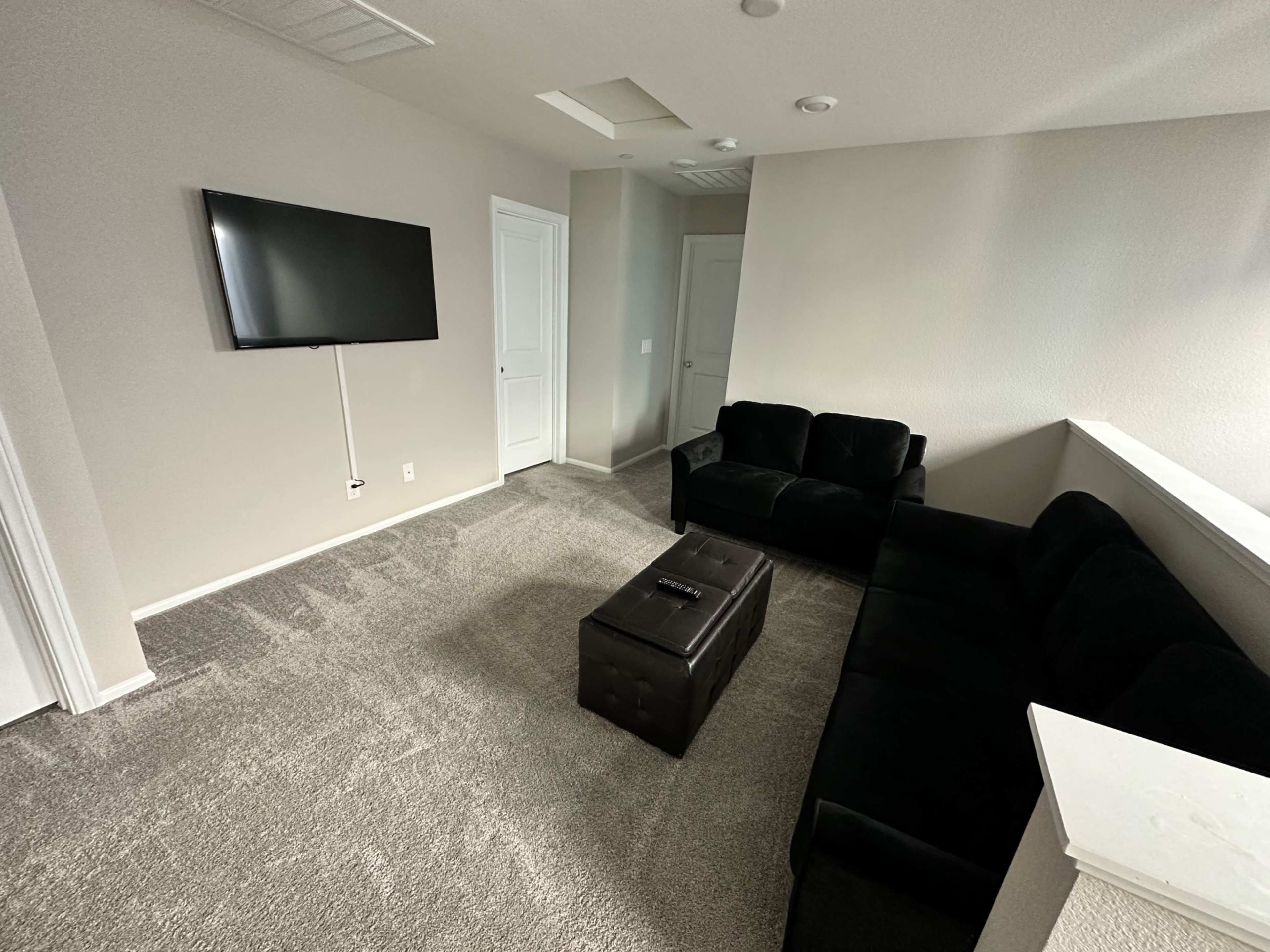 The image shows a small living room with a black couch, a coffee table, a television mounted on the wall, and light-colored walls and carpet.