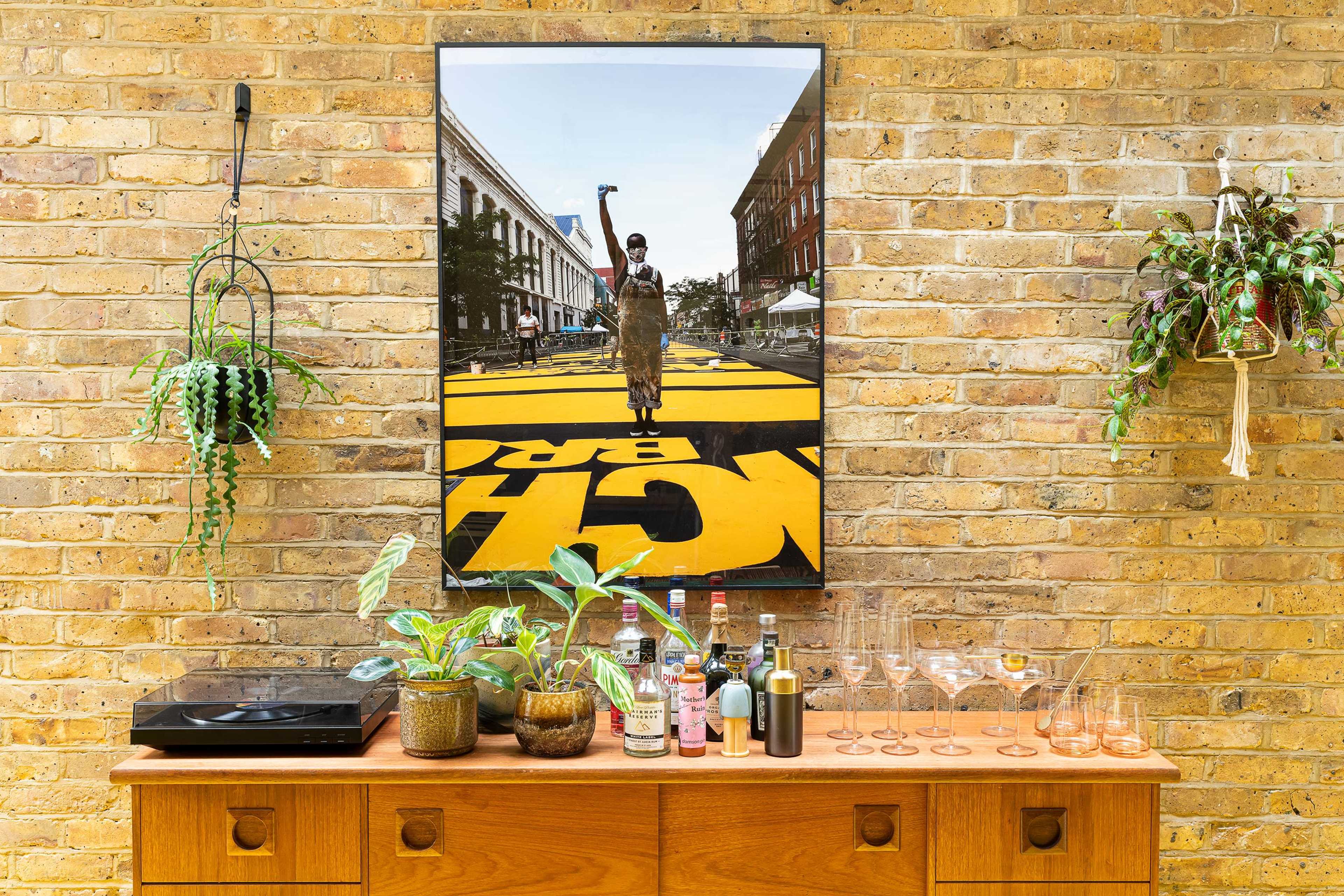 A wooden sideboard with various plants, bottles, and glasses sits against a brick wall, featuring a large framed photograph of a person walking on a street marked with yellow paint.