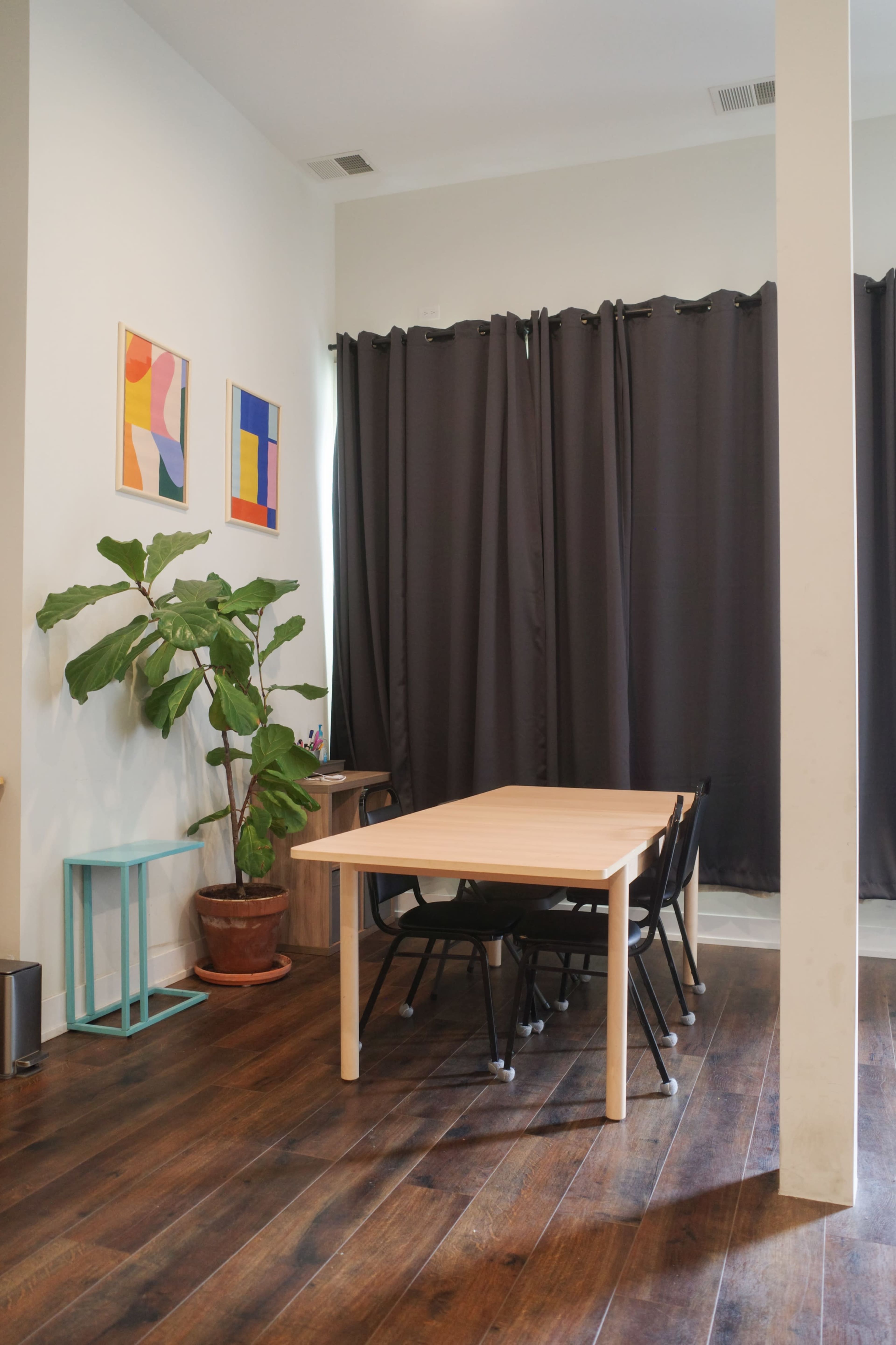 The image shows a room with a wooden table and black chairs, a large potted plant, and artwork on the wall, complemented by dark curtains.