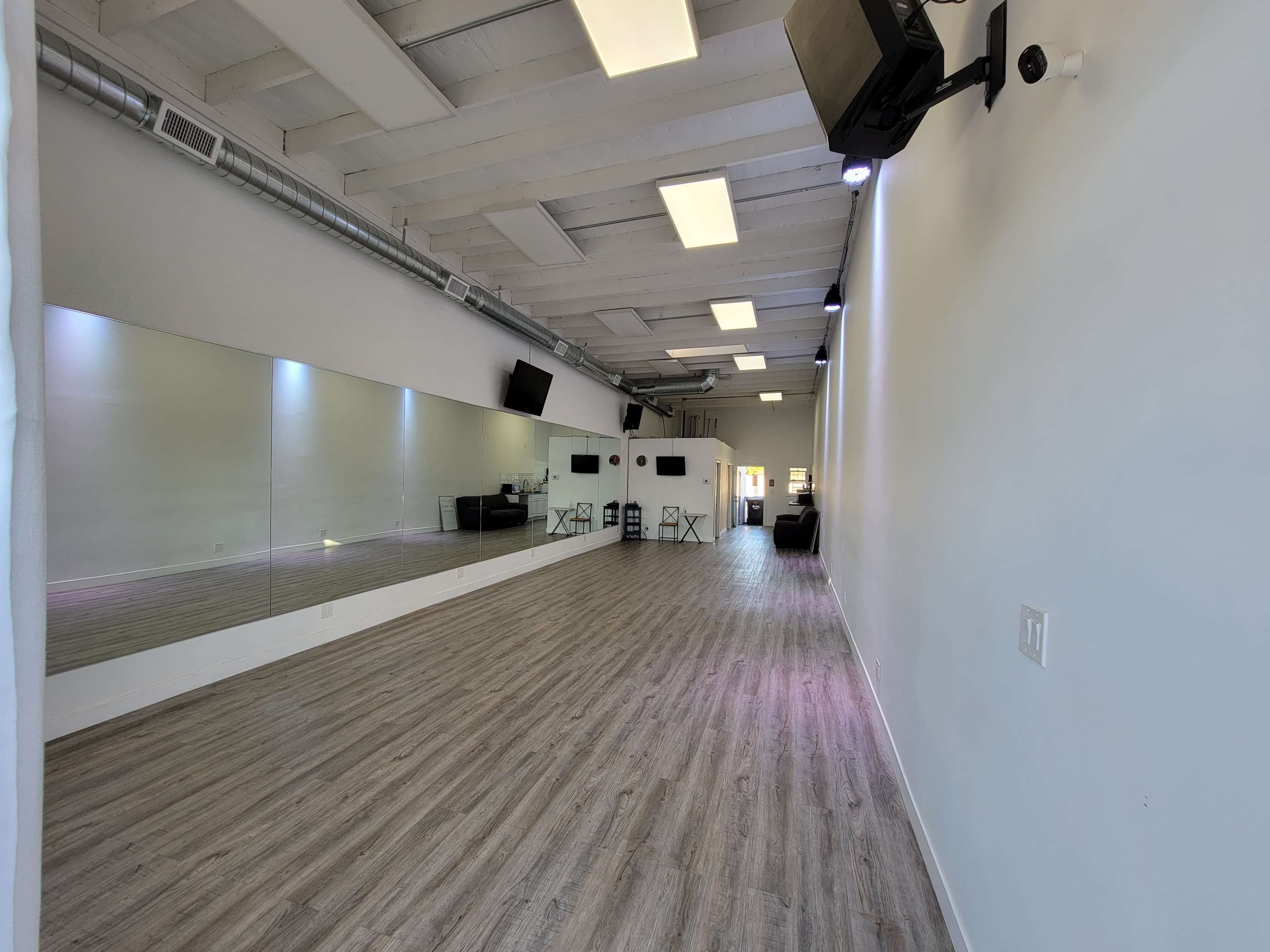 The image shows a spacious dance studio with wooden flooring, a large mirror on one wall, and overhead lights illuminating the area.