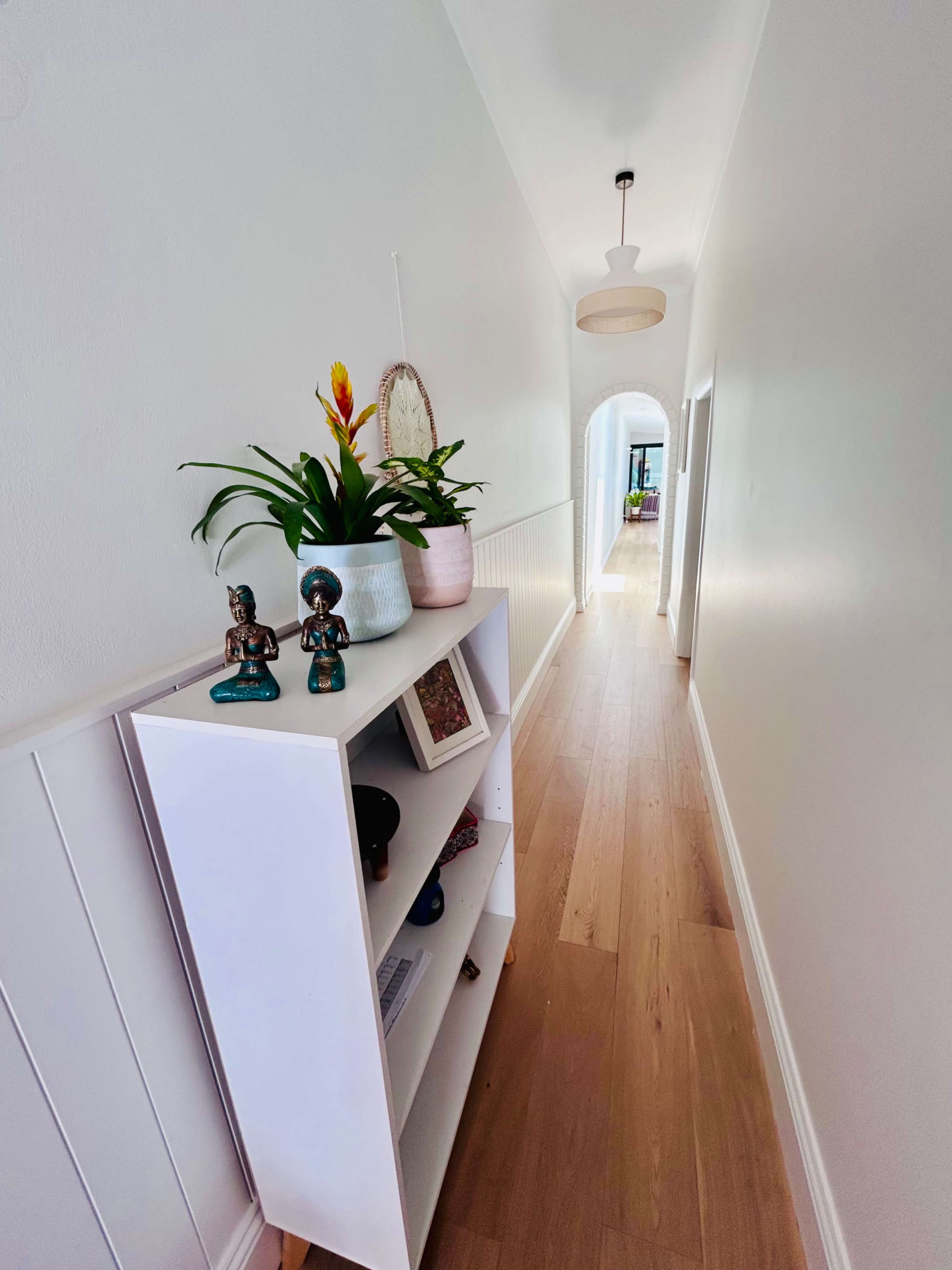 The image shows a narrow hallway with light wooden flooring, a white wall, and a decorative shelf on the left side featuring plants and small sculptures.