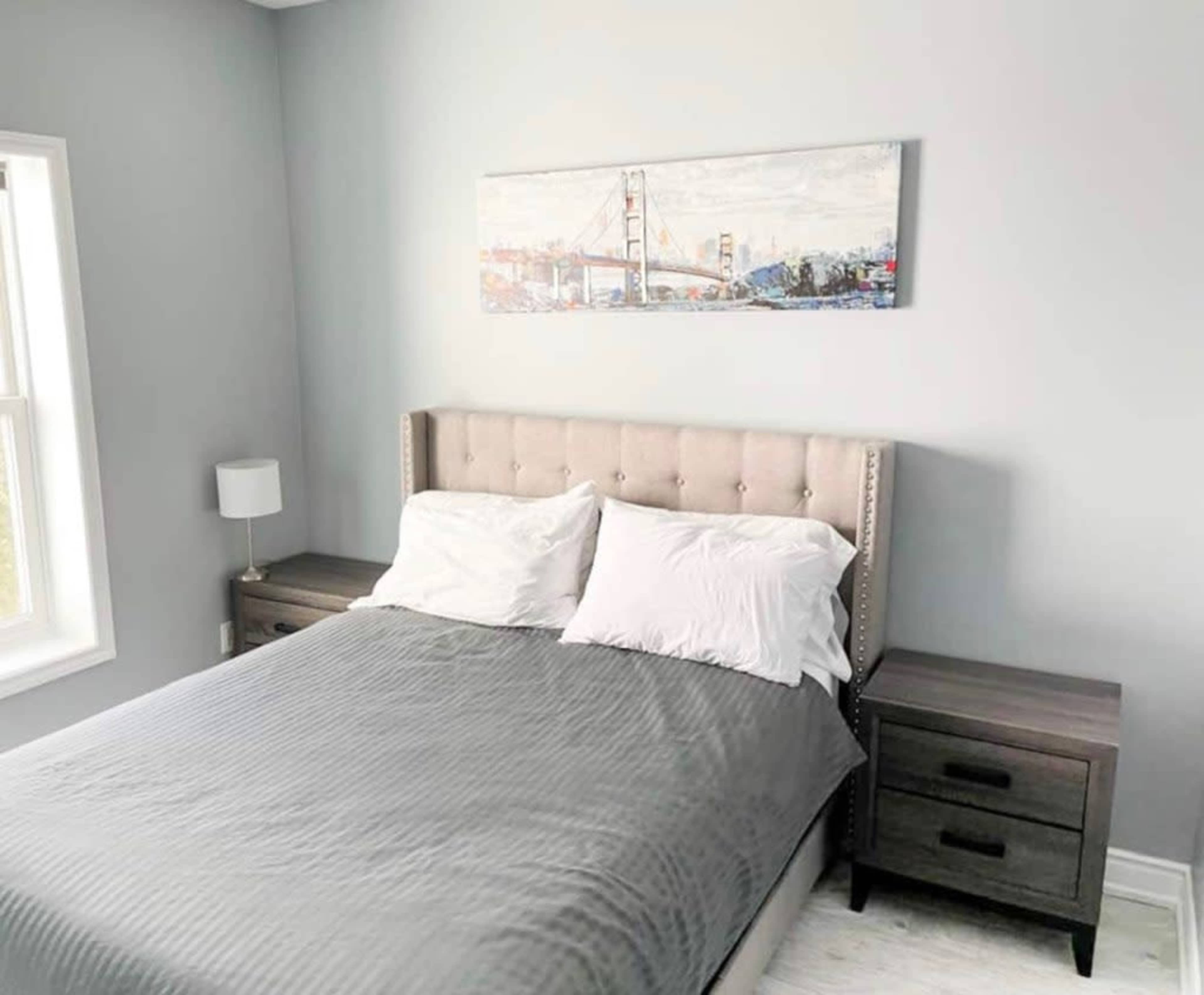 The room features a bed with a tufted headboard, flanked by two nightstands, and a landscape painting above the headboard.