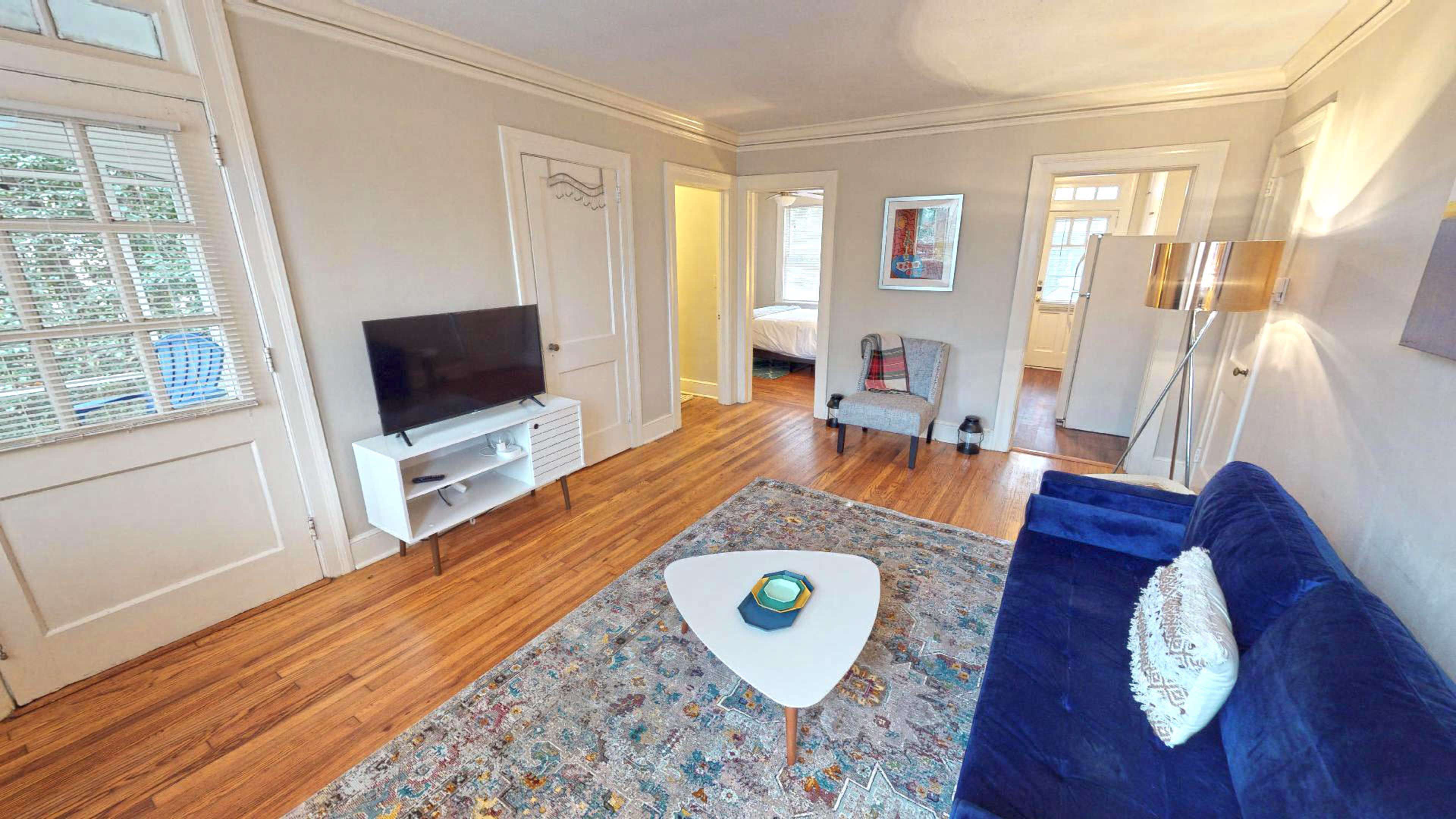 The image shows a living room with a blue sofa, a small coffee table, a TV stand with a television, and wooden floors, accented by a decorative rug.