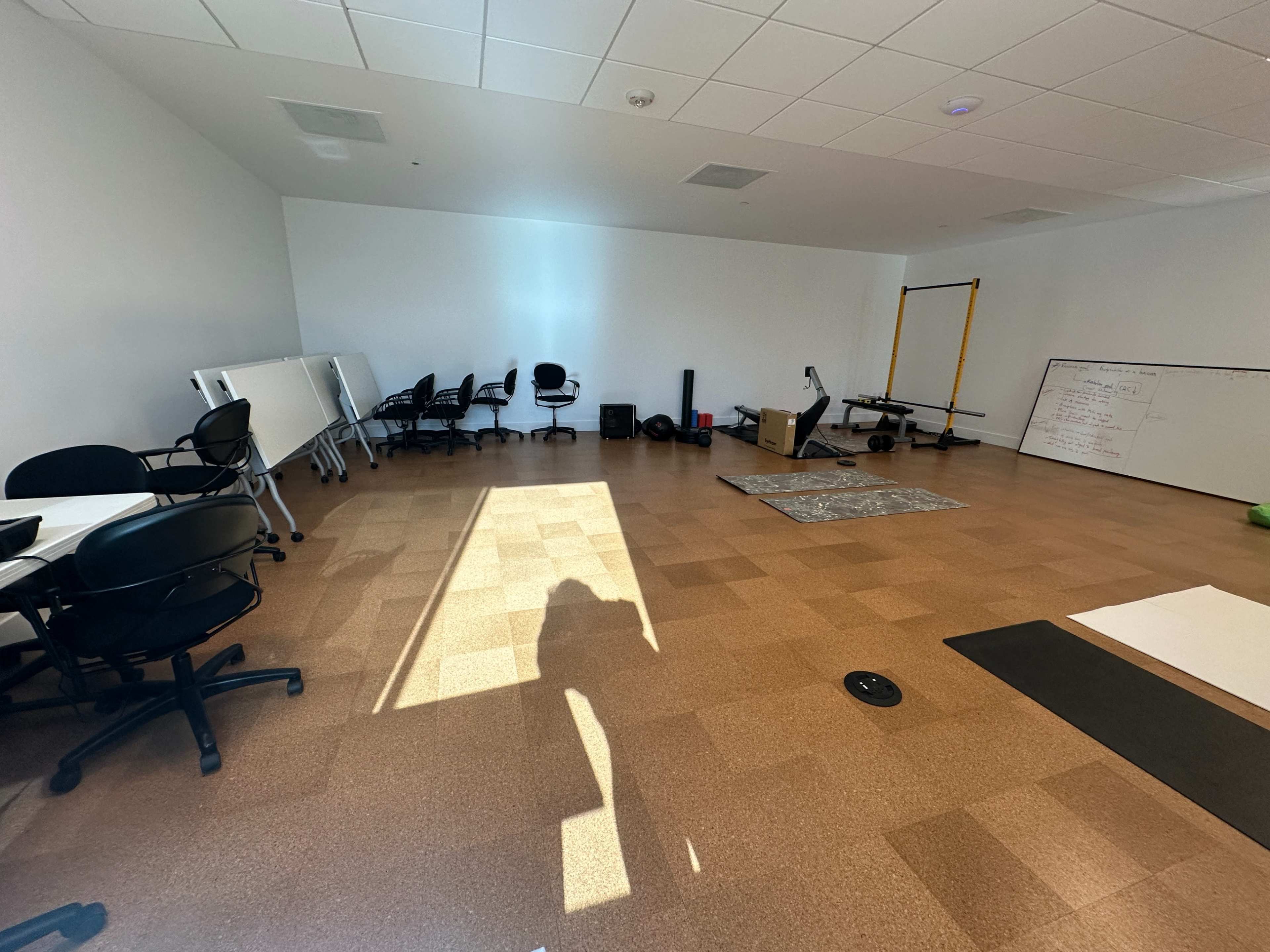 The image shows a spacious, empty room with several chairs, tables, a whiteboard, and exercise equipment scattered throughout.