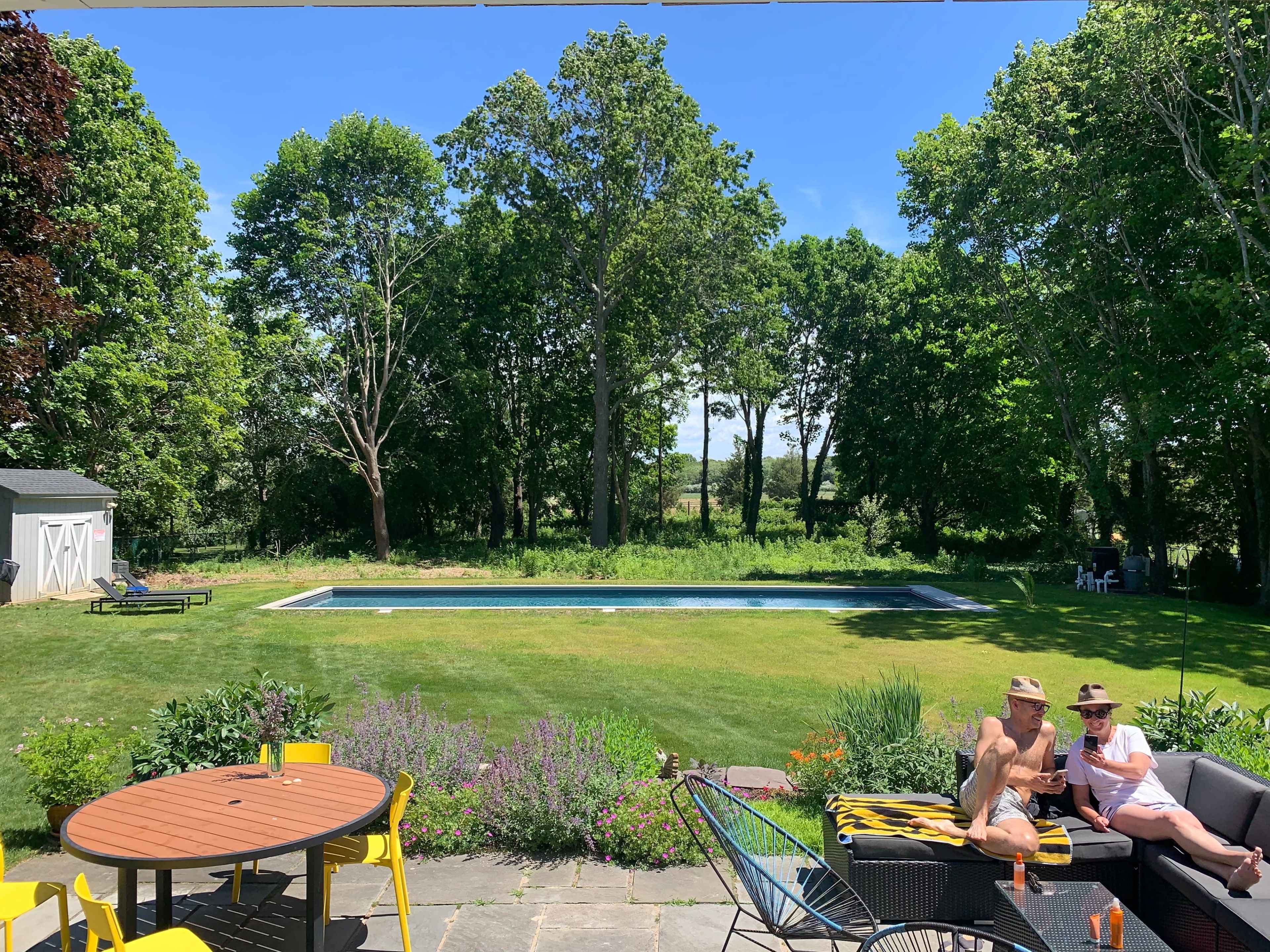 A group of people relaxes near a swimming pool surrounded by trees and a landscaped garden.