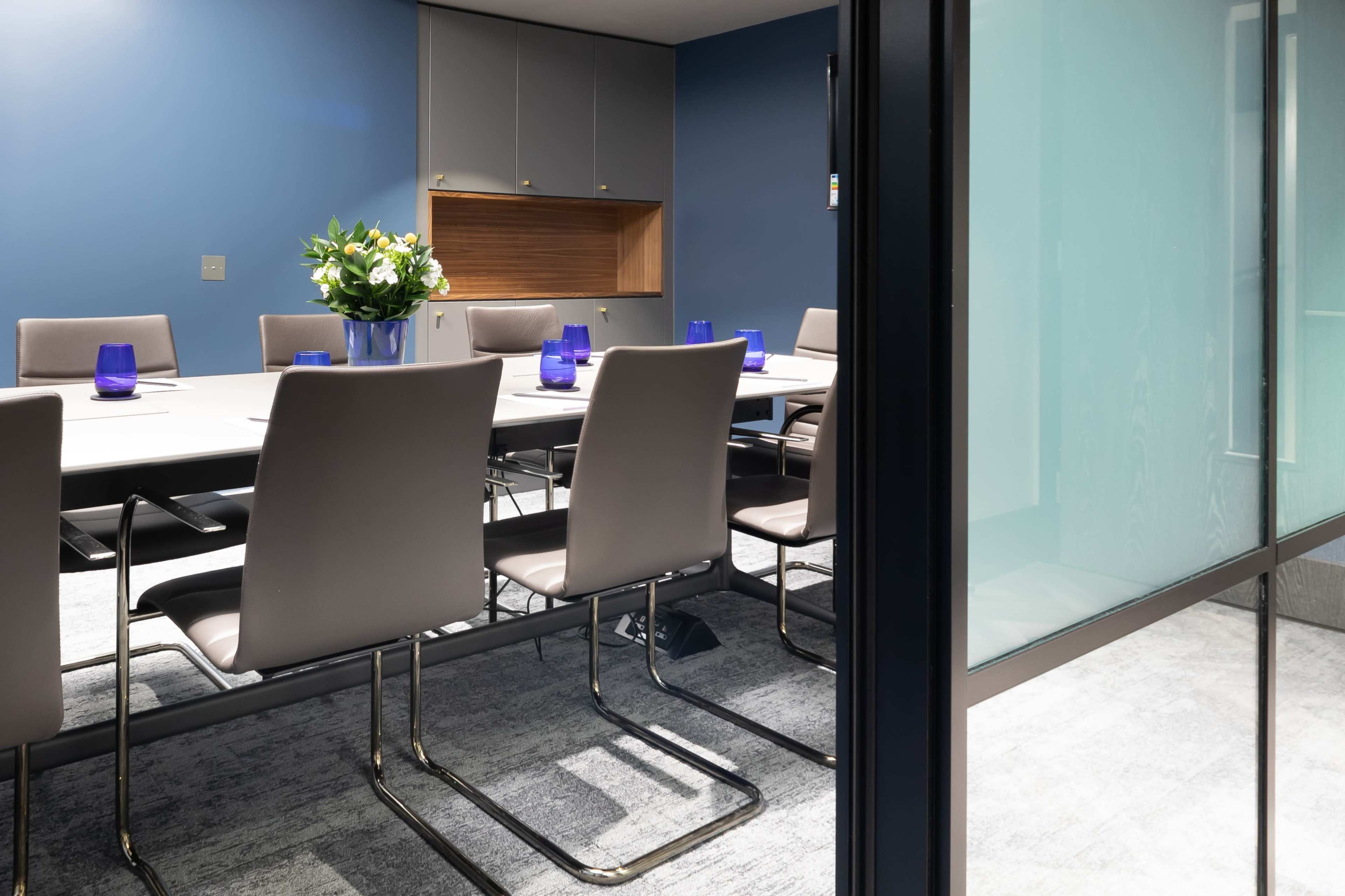 A modern conference room features a long table surrounded by gray chairs, with a blue accent wall and a floral centerpiece.