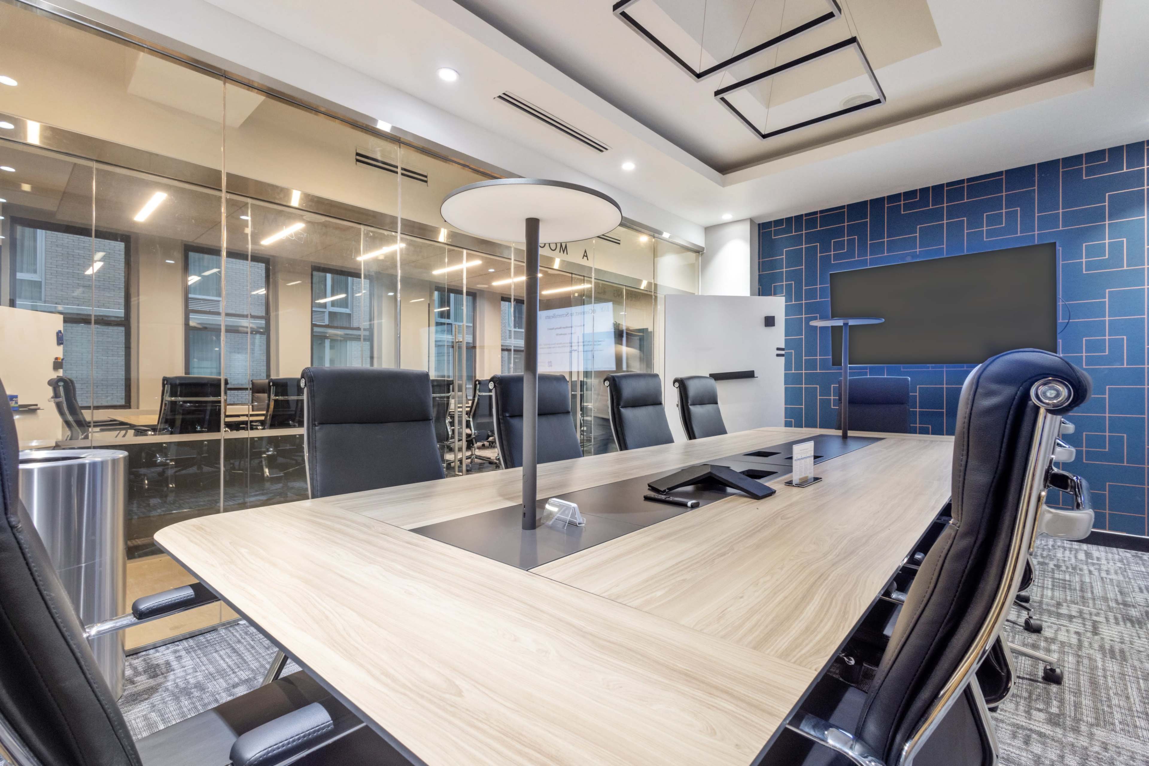 A modern conference room features a large wooden table surrounded by black executive chairs, with glass walls and a digital screen on the blue patterned wall.