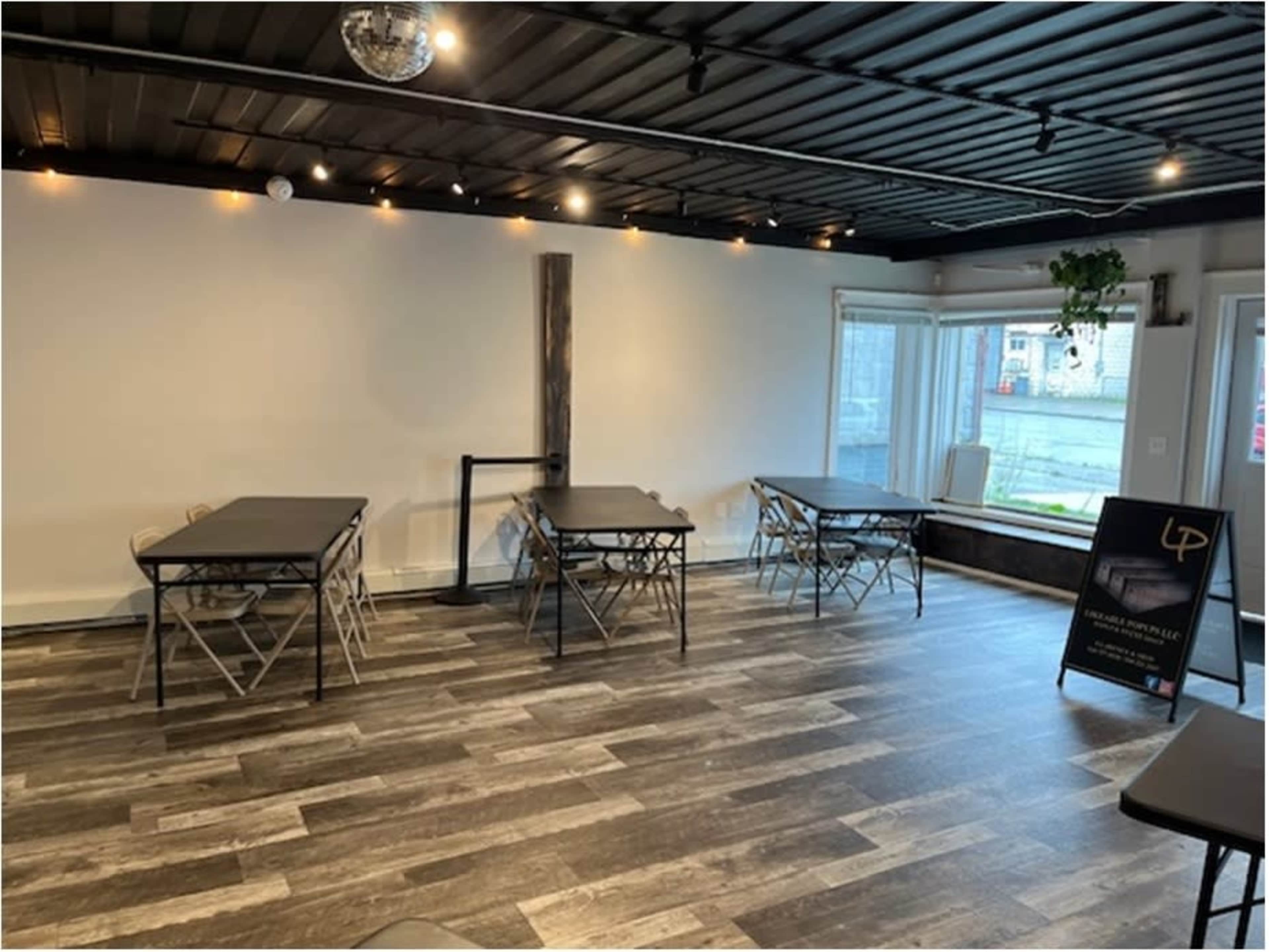 A sparsely furnished interior space with several folding tables arranged on a wooden floor and large windows letting in natural light.
