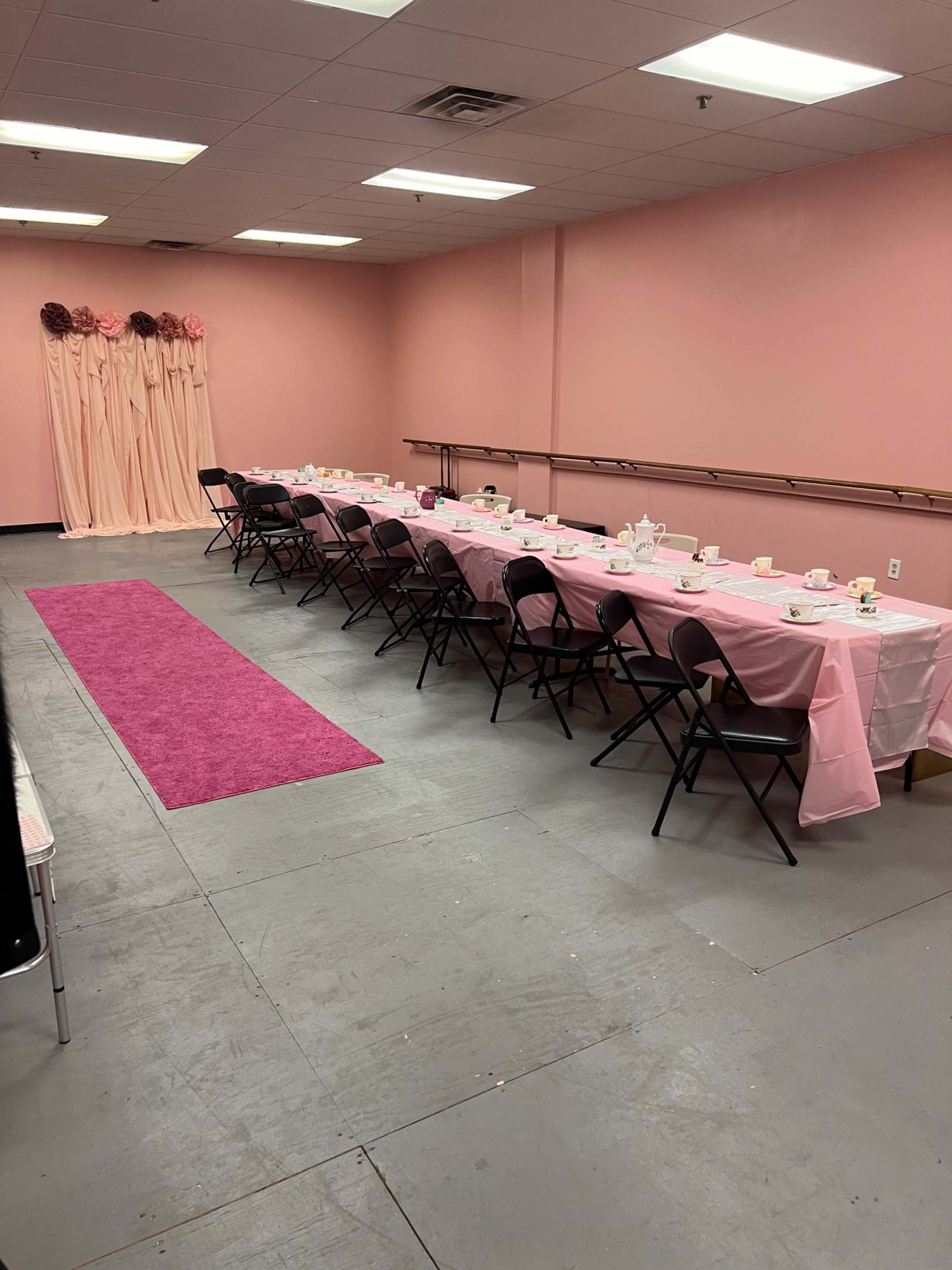 A pink-themed banquet room set with a long table adorned with a pink tablecloth, black chairs, and tea service items, accompanied by a pink carpet and decorative backdrop.