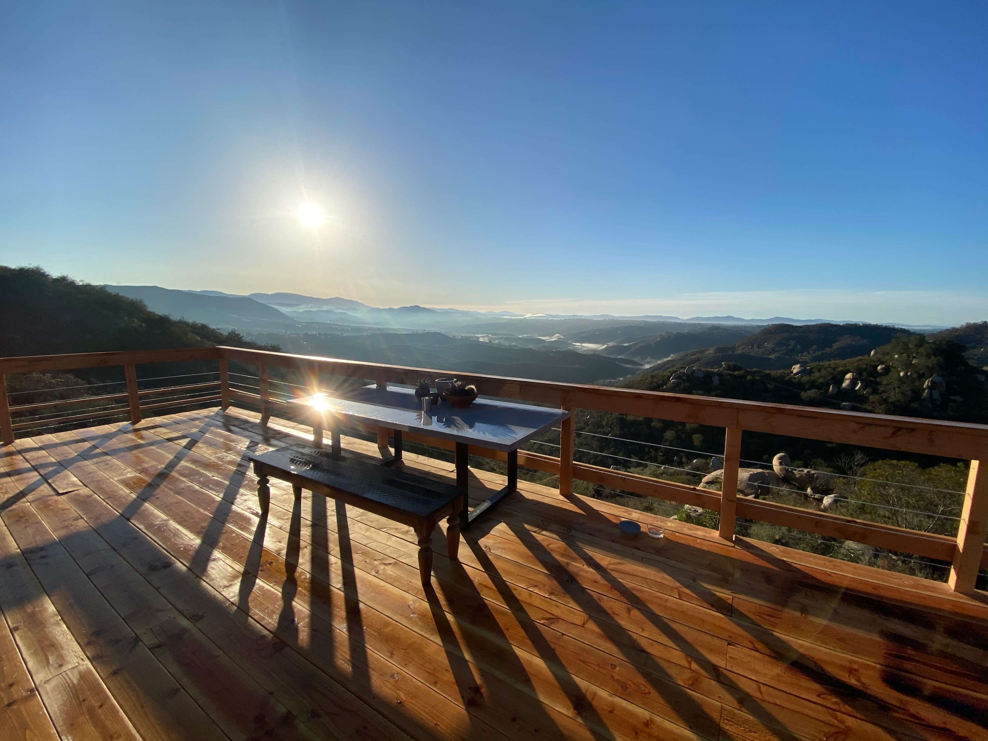 A wooden deck overlooks a mountainous landscape with the sun rising in the background.