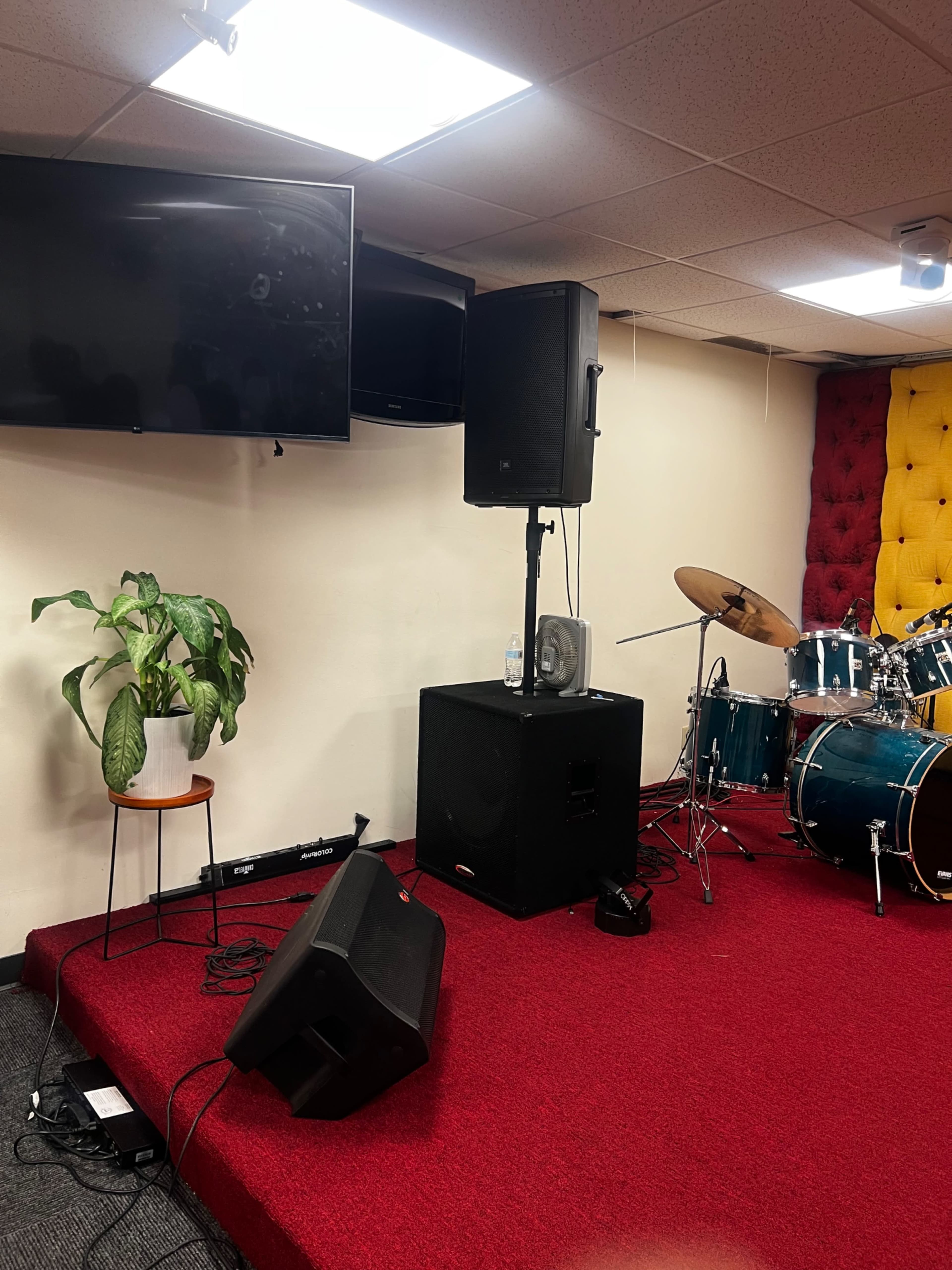 The image shows a small music performance space with a red carpeted stage, a drum set, a speaker, and a potted plant on a stool.