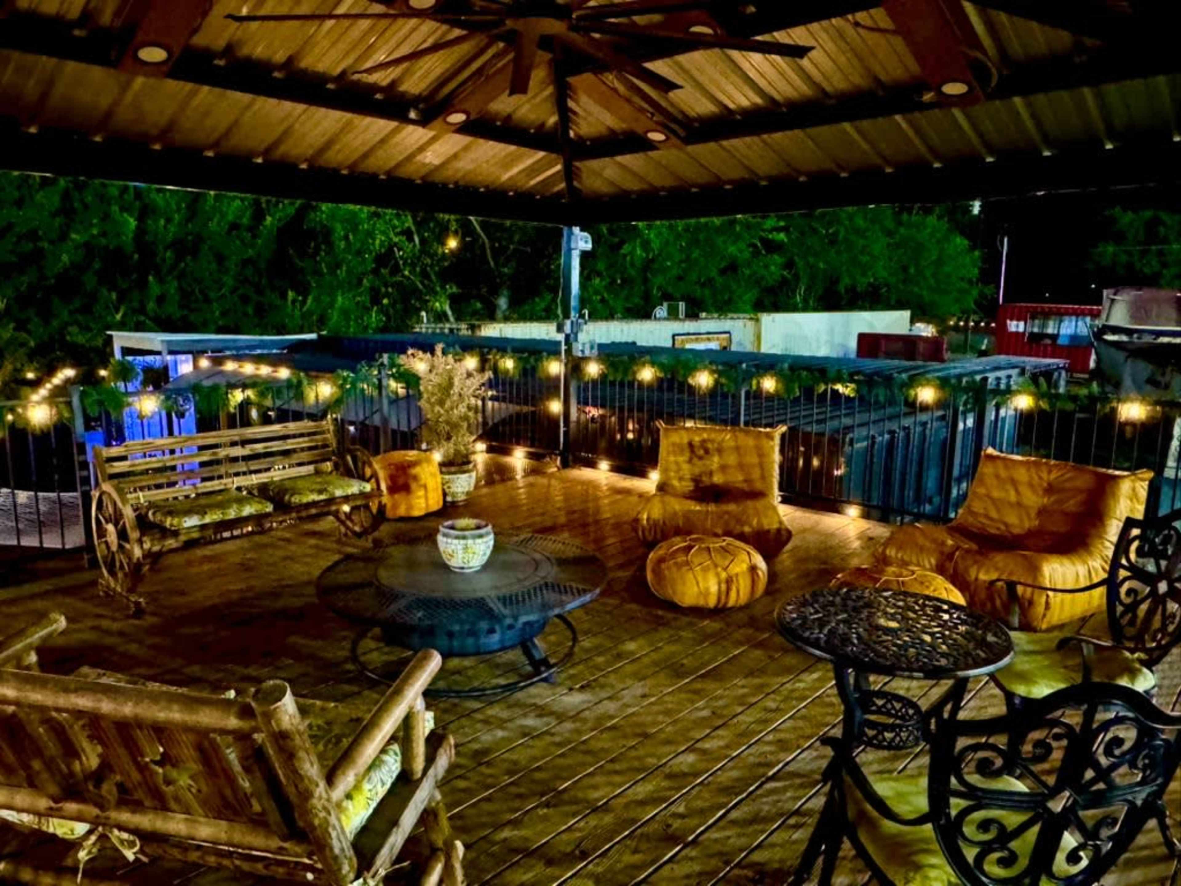The image shows a cozy outdoor seating area with a wooden deck, featuring a round table, various chairs, and ambient lighting strung around the space.