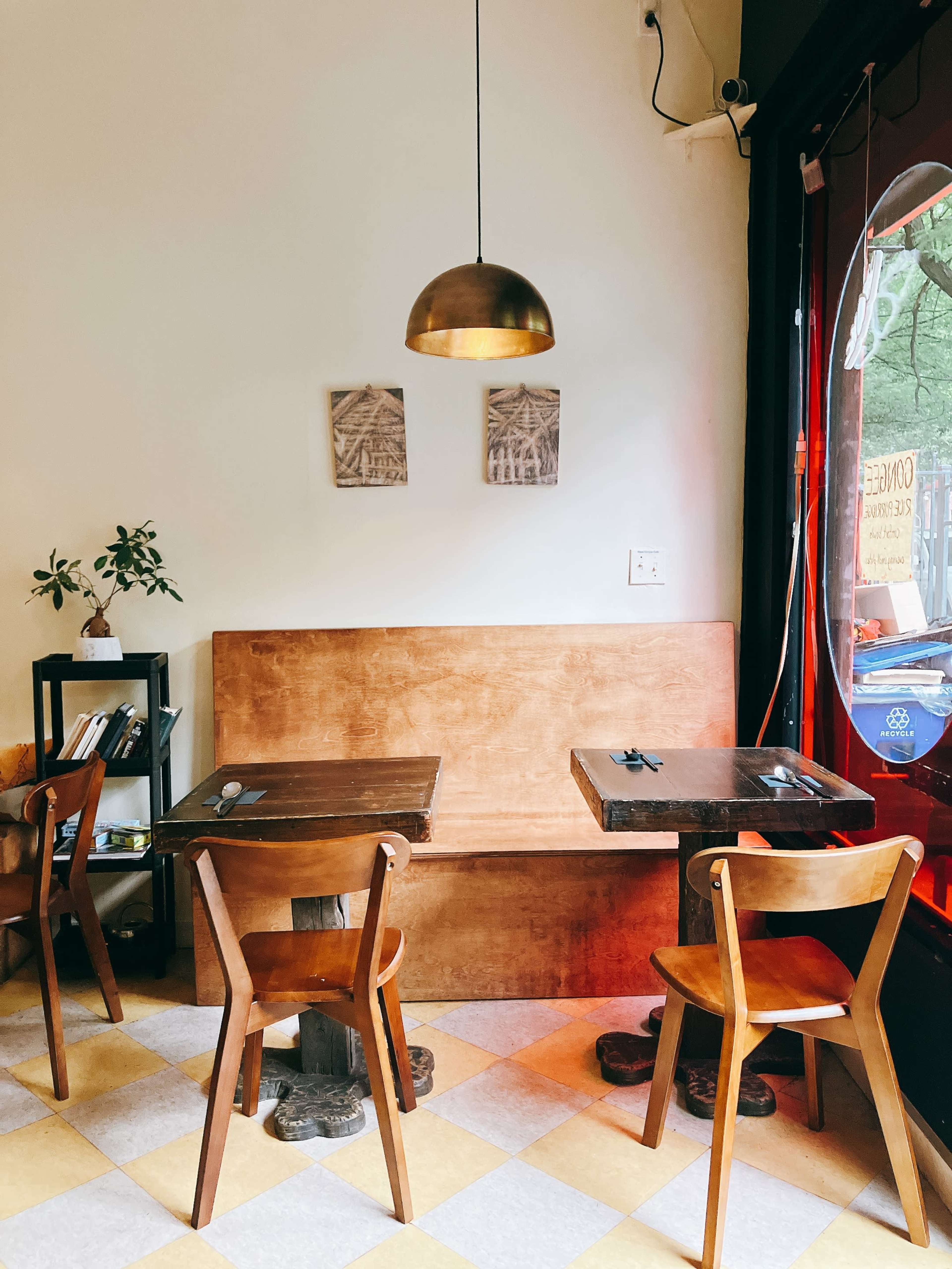 The image shows a small cafe interior featuring wooden tables and chairs, a pendant light hanging from the ceiling, and a window with a view of the outdoors.