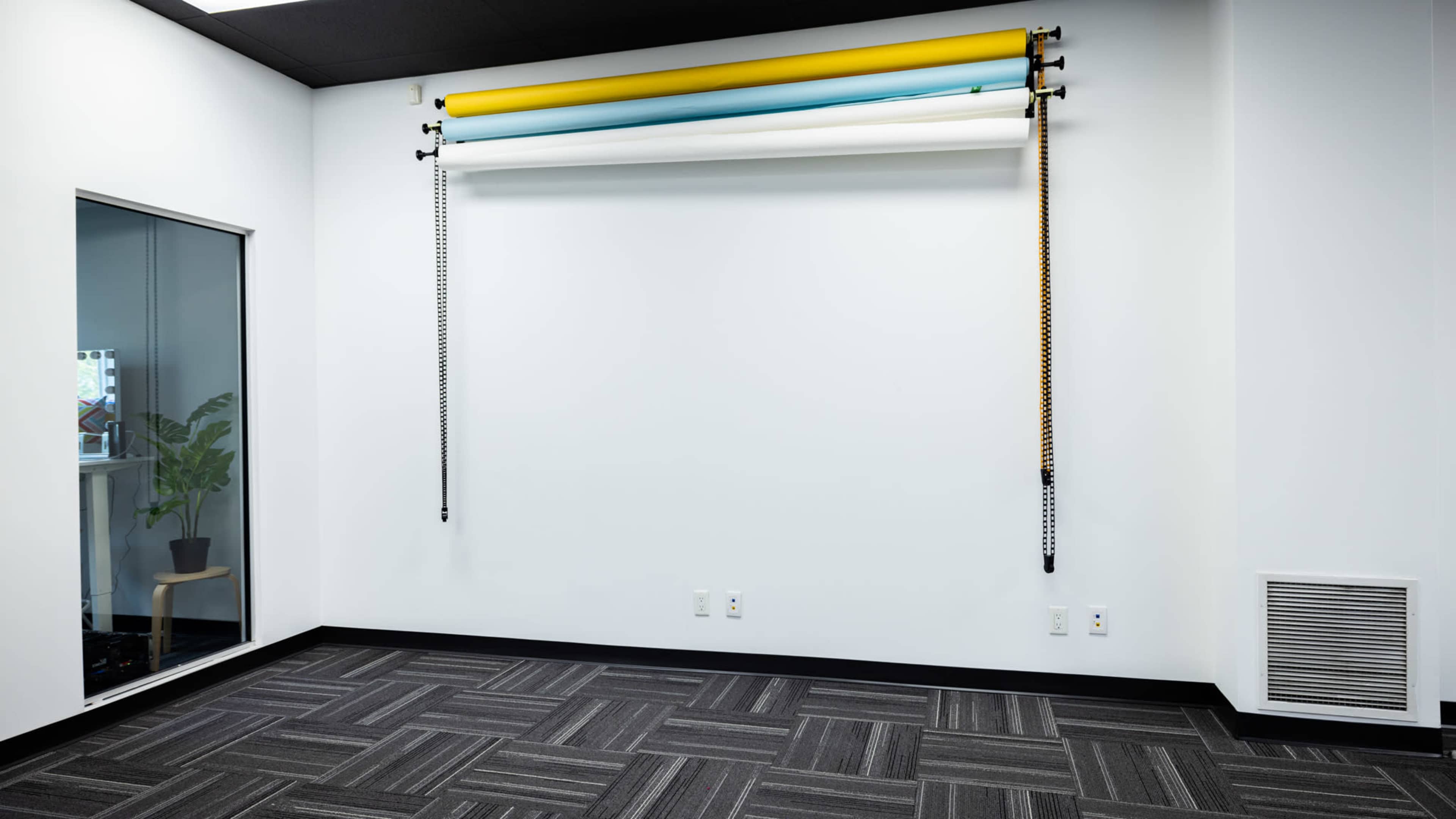 A sparsely furnished room with a white wall featuring rolls of colored backdrop paper mounted above, and a large window revealing a plant in the adjacent space.