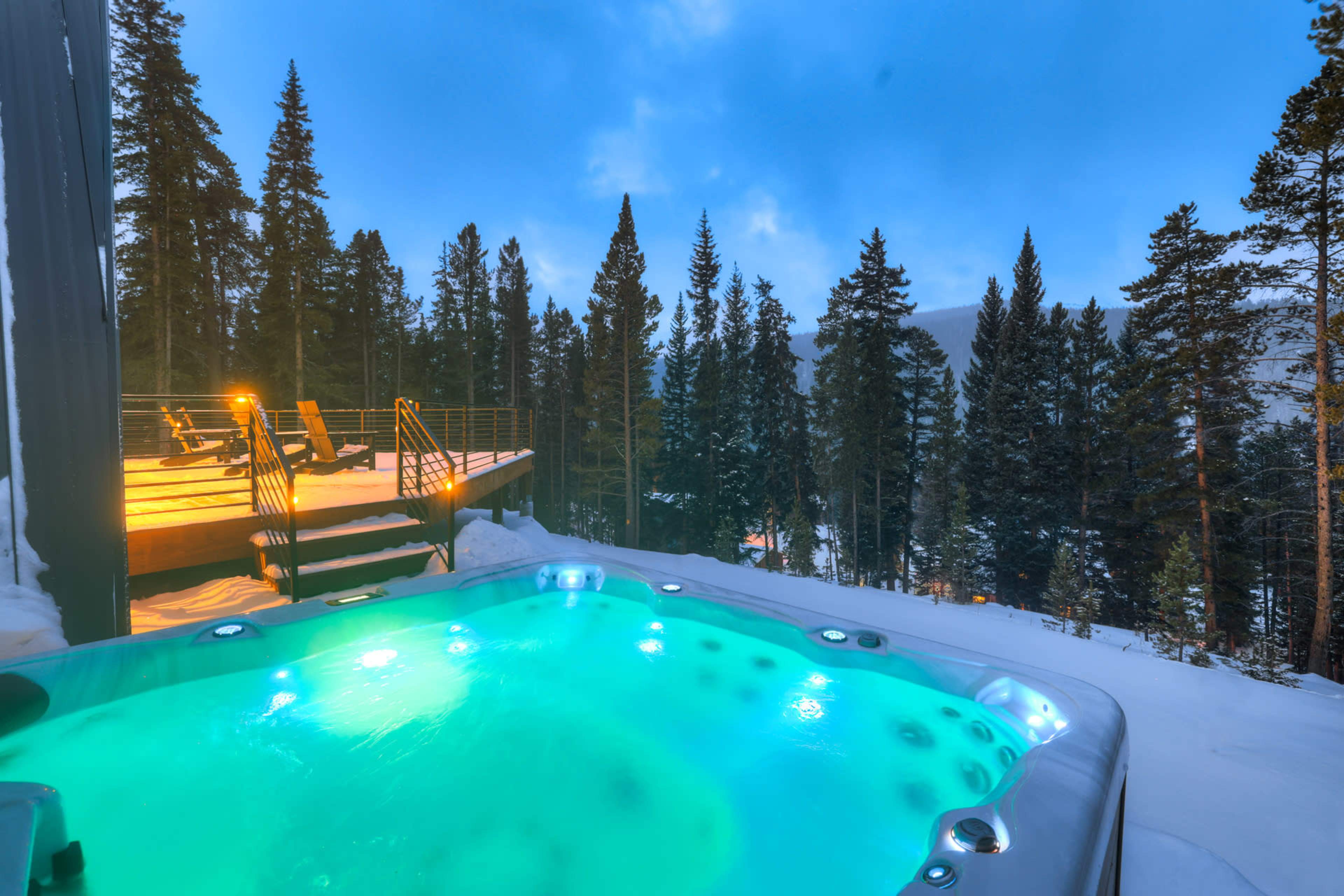 A hot tub with illuminated water sits on a snowy deck surrounded by tall pine trees under a blue sky.