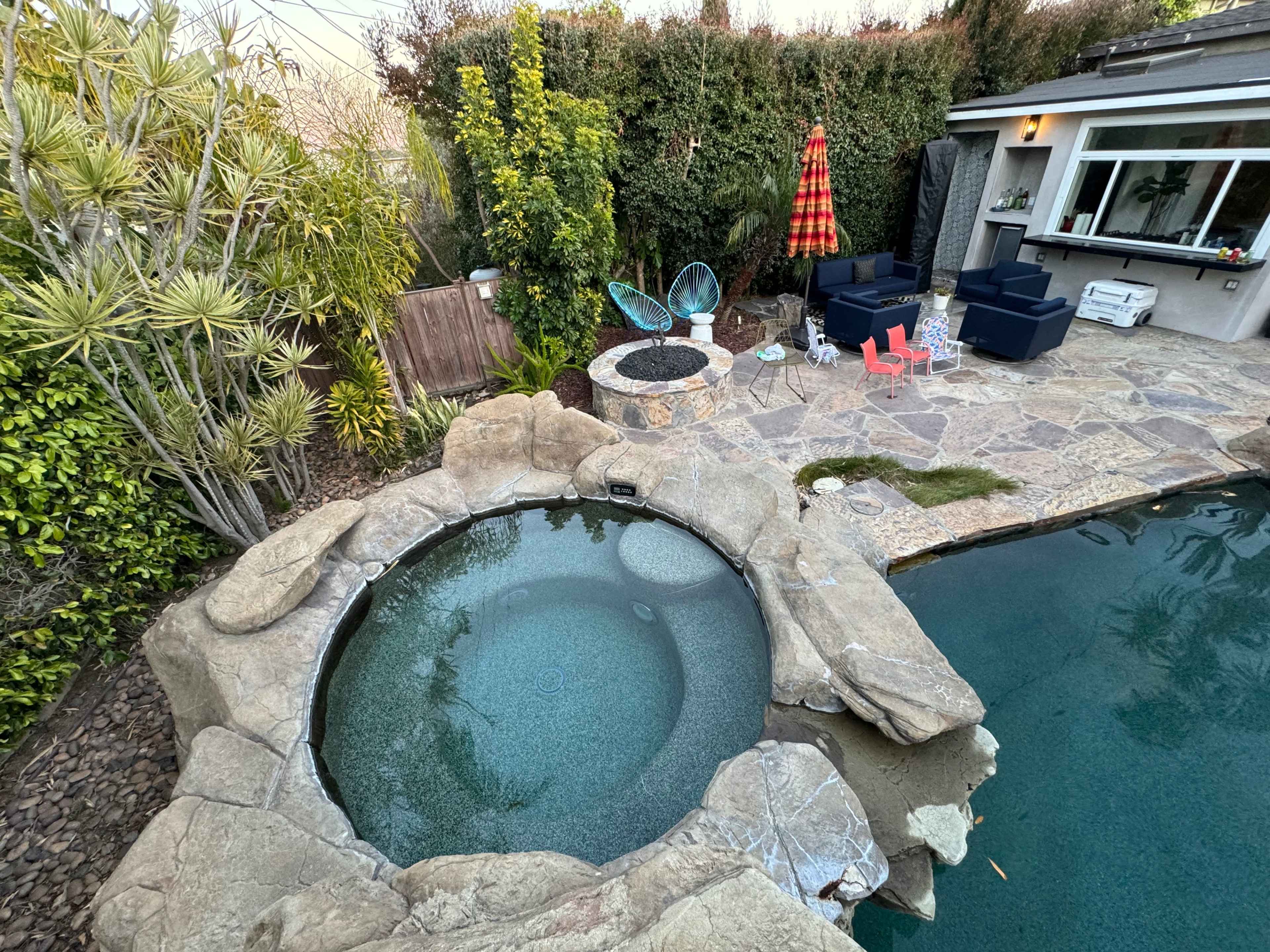 A backyard with a stone hot tub and a swimming pool, surrounded by greenery and outdoor seating.
