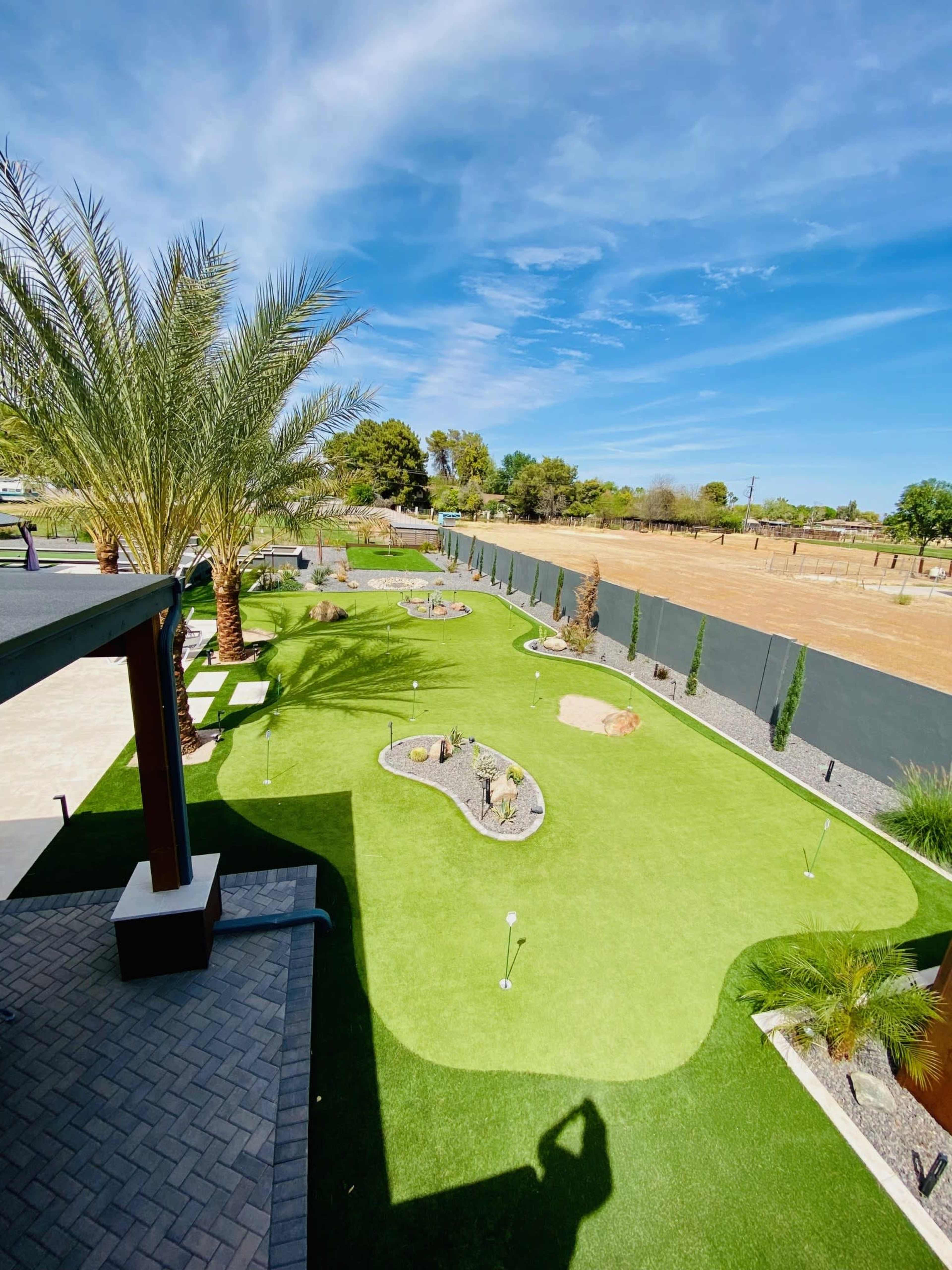 The image shows a landscaped backyard featuring a putting green surrounded by ornamental plants and decorative stones under a clear blue sky.