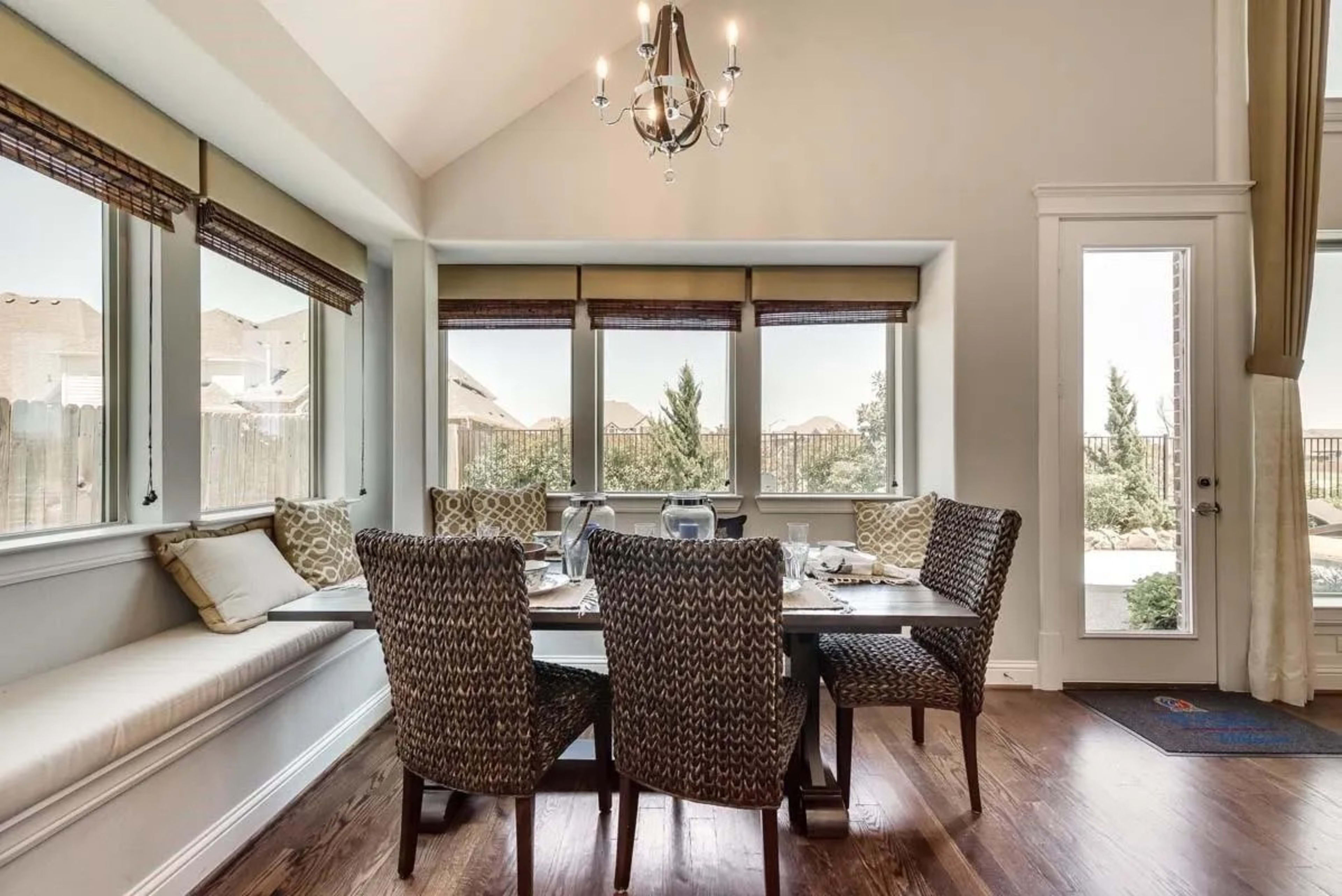 A dining area features a table surrounded by patterned chairs, with large windows allowing natural light and a view of the outdoors.