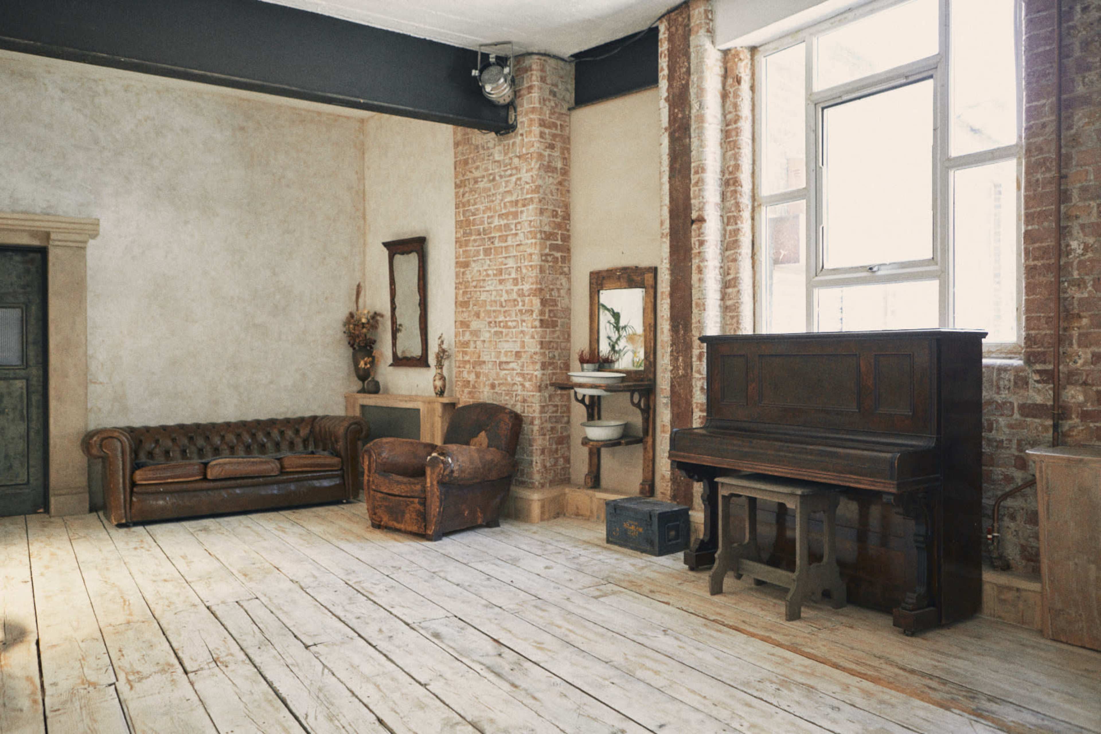 The room features a wooden floor, exposed brick walls, a vintage leather sofa, an antique armchair, and a piano.